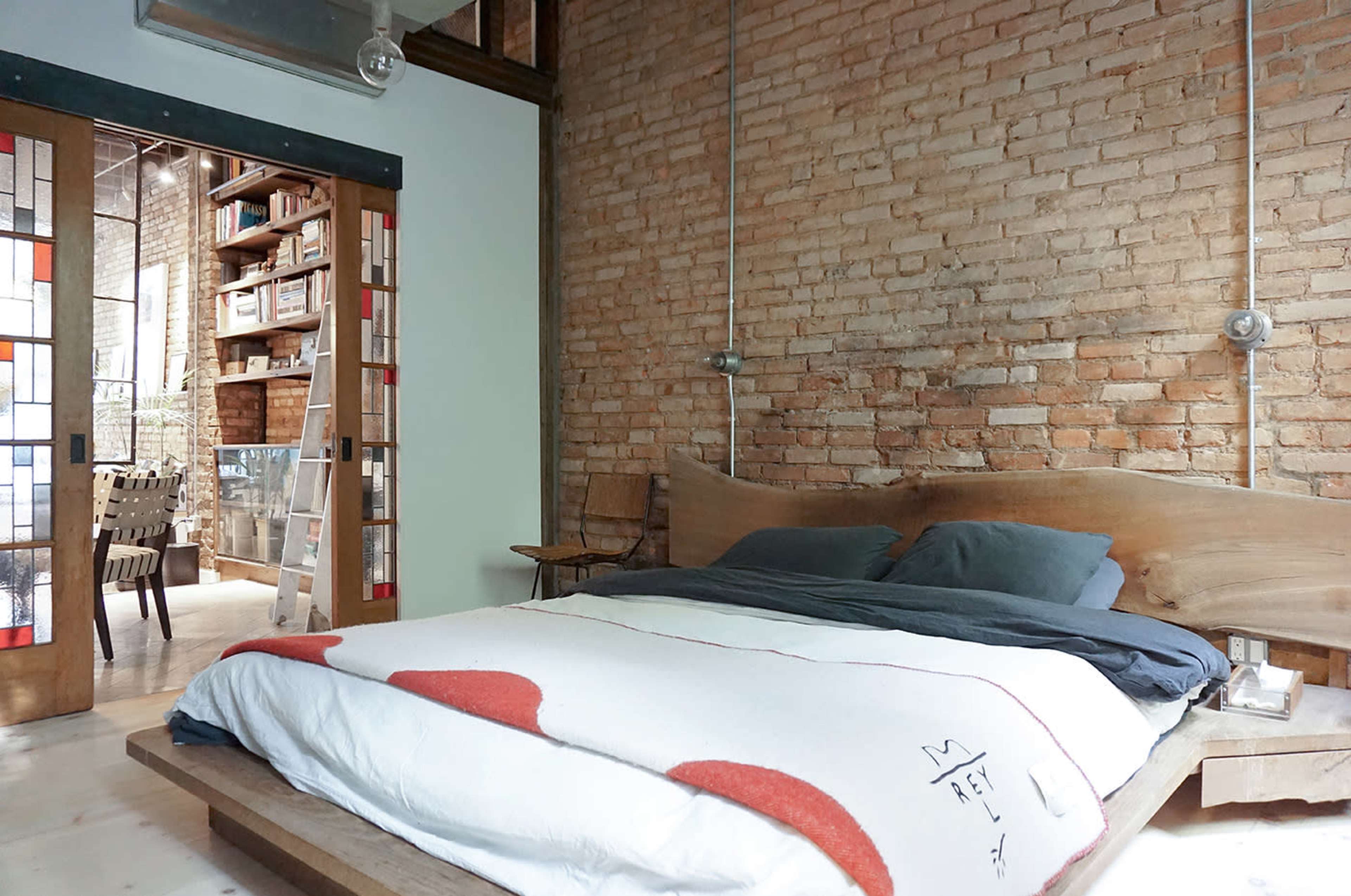 A cozy bedroom with a wooden bed and exposed brick walls, leading to a dining area visible through a set of glass-paneled doors.