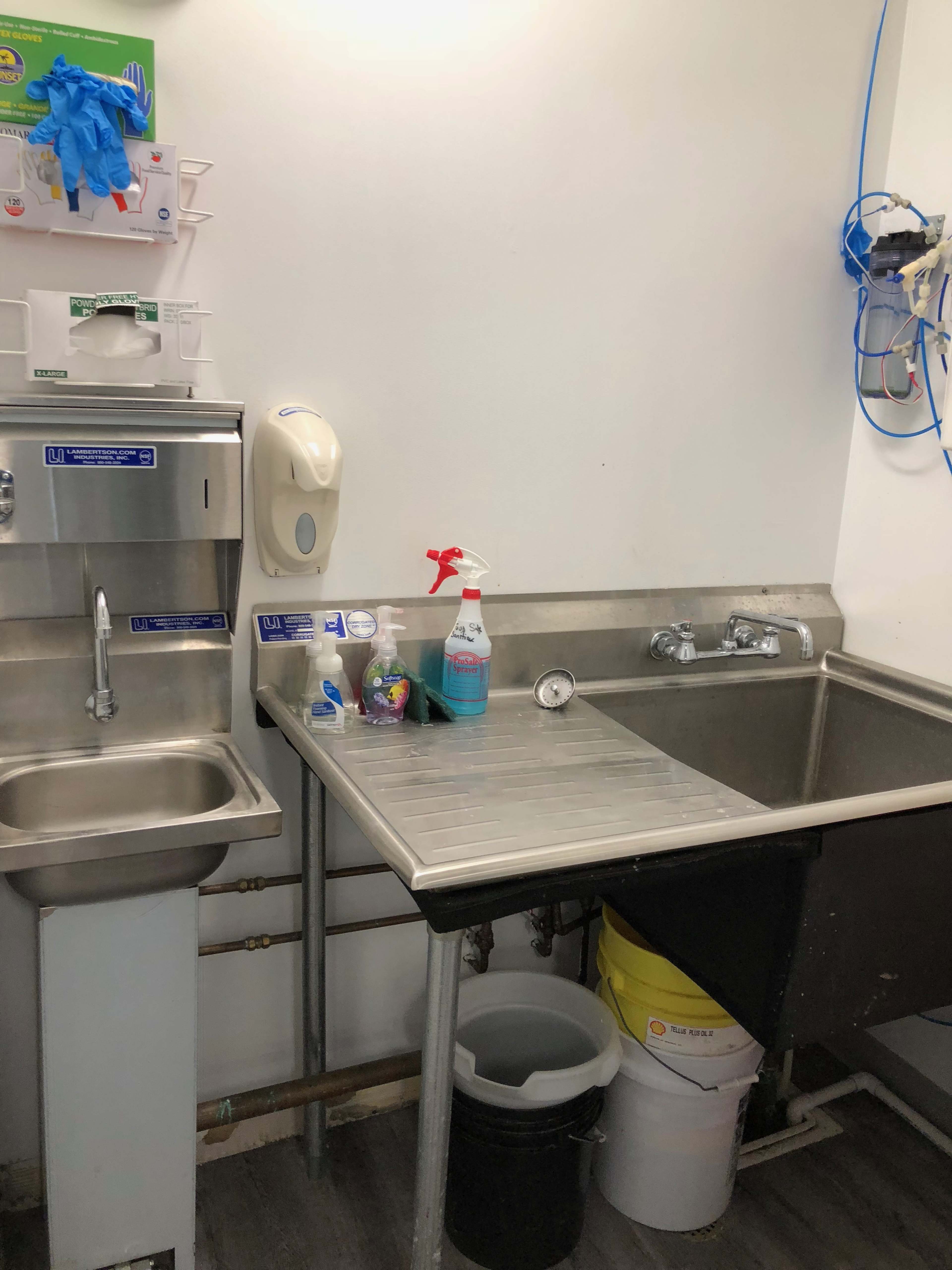 The image shows a small commercial kitchen area with a stainless steel sink, a spray bottle, cleaning supplies, and a waste container.