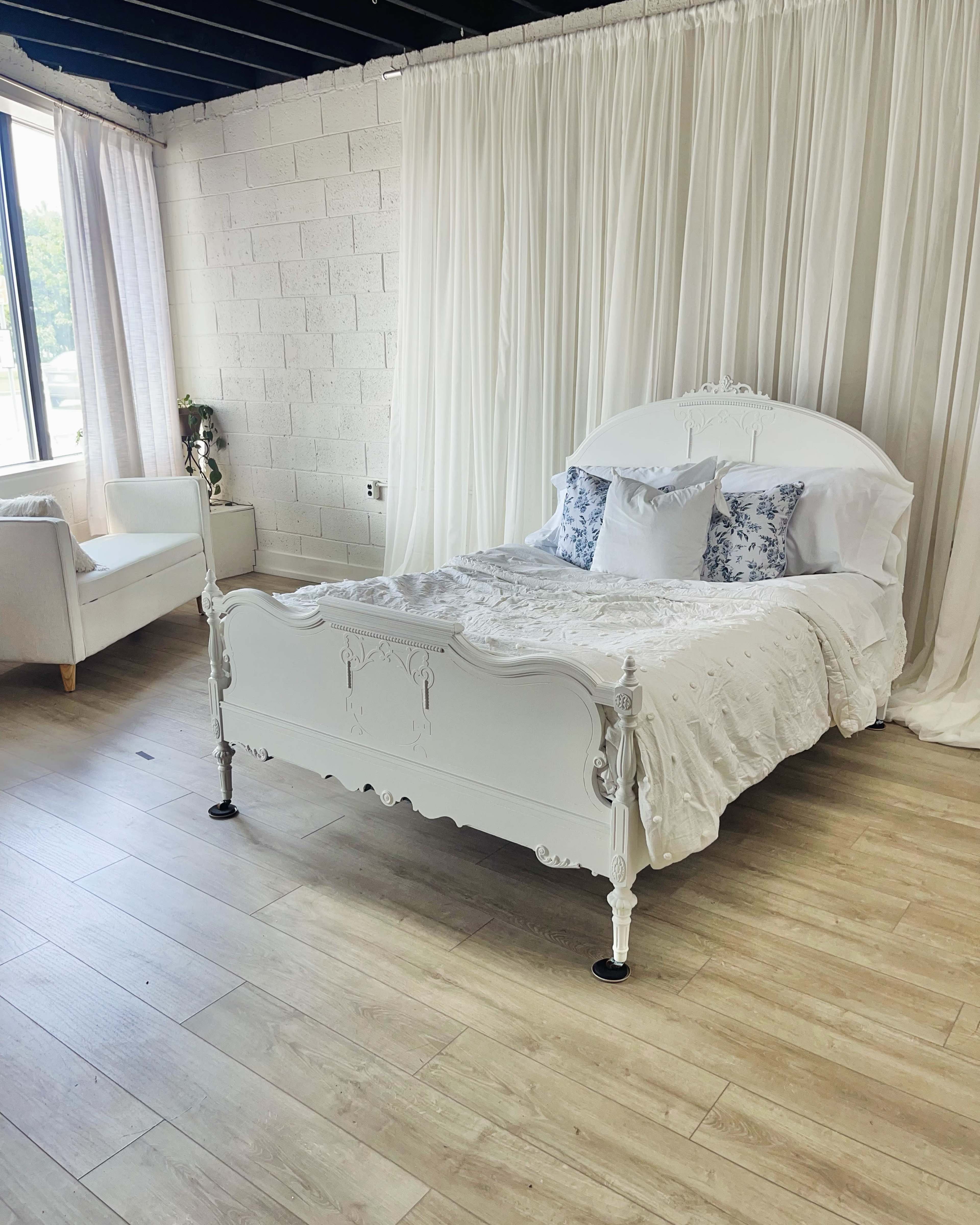 The image shows a white, ornate bed with a light-colored bedding set positioned in a room with large windows and sheer curtains.