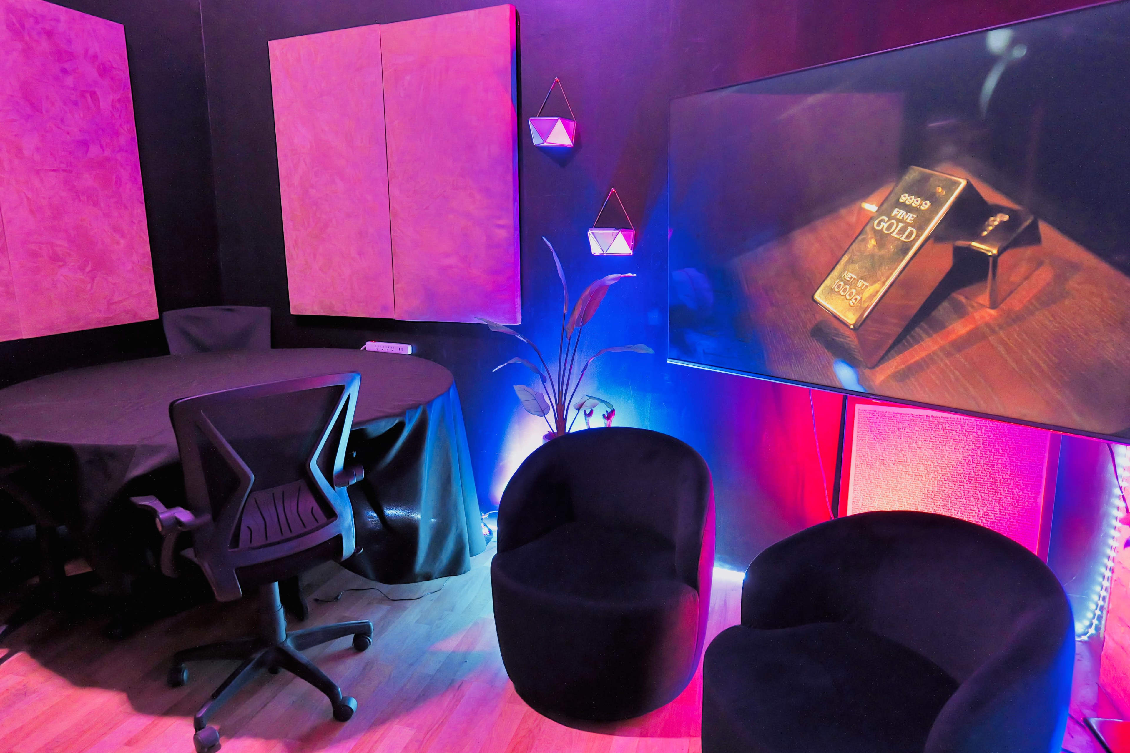 A modern meeting room featuring a round table, a swivel chair, and two black lounge chairs, illuminated by colored lights and a large screen displaying an object.