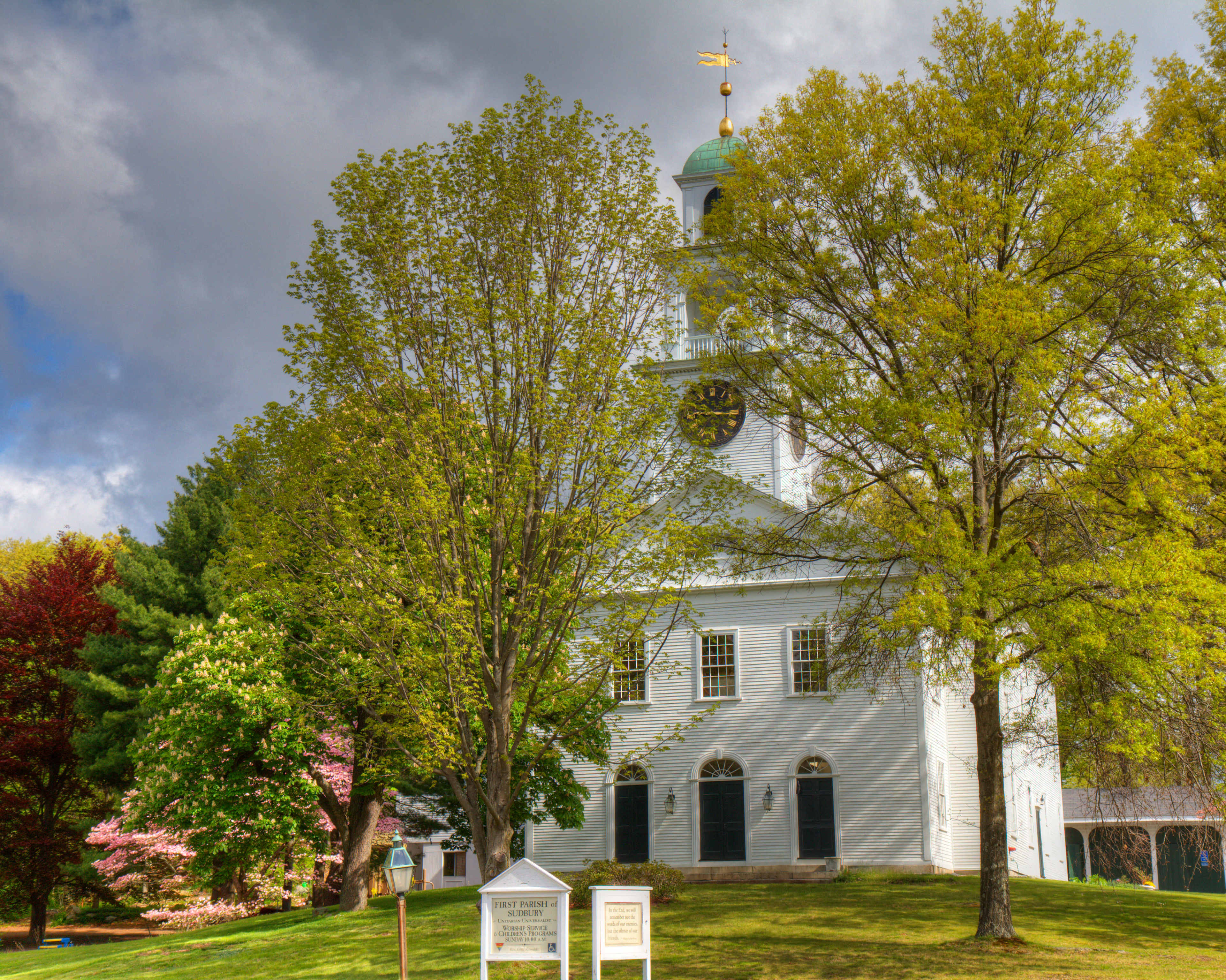 A white church with a green dome and clock tower is surrounded by several trees and a grassy area under a partly cloudy sky.
