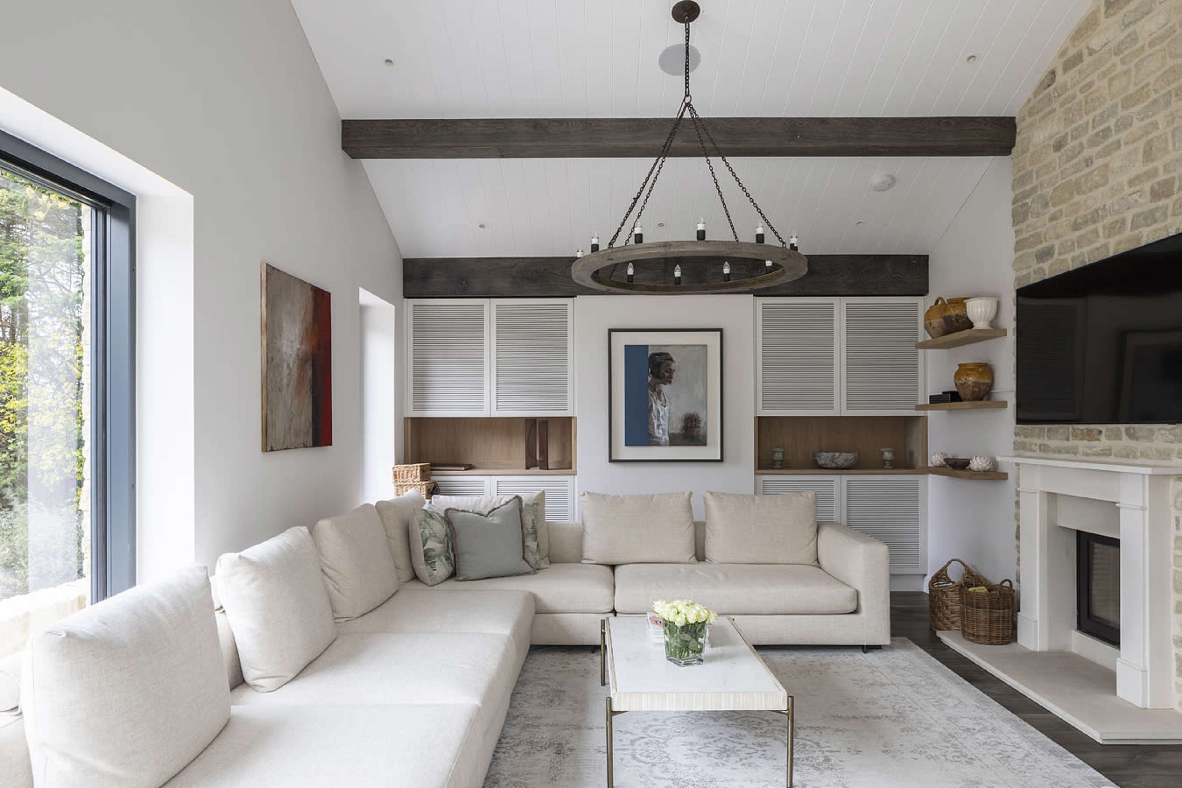 A bright living room features a large light-colored sectional sofa, a round chandelier, and a stone wall with a mounted television.