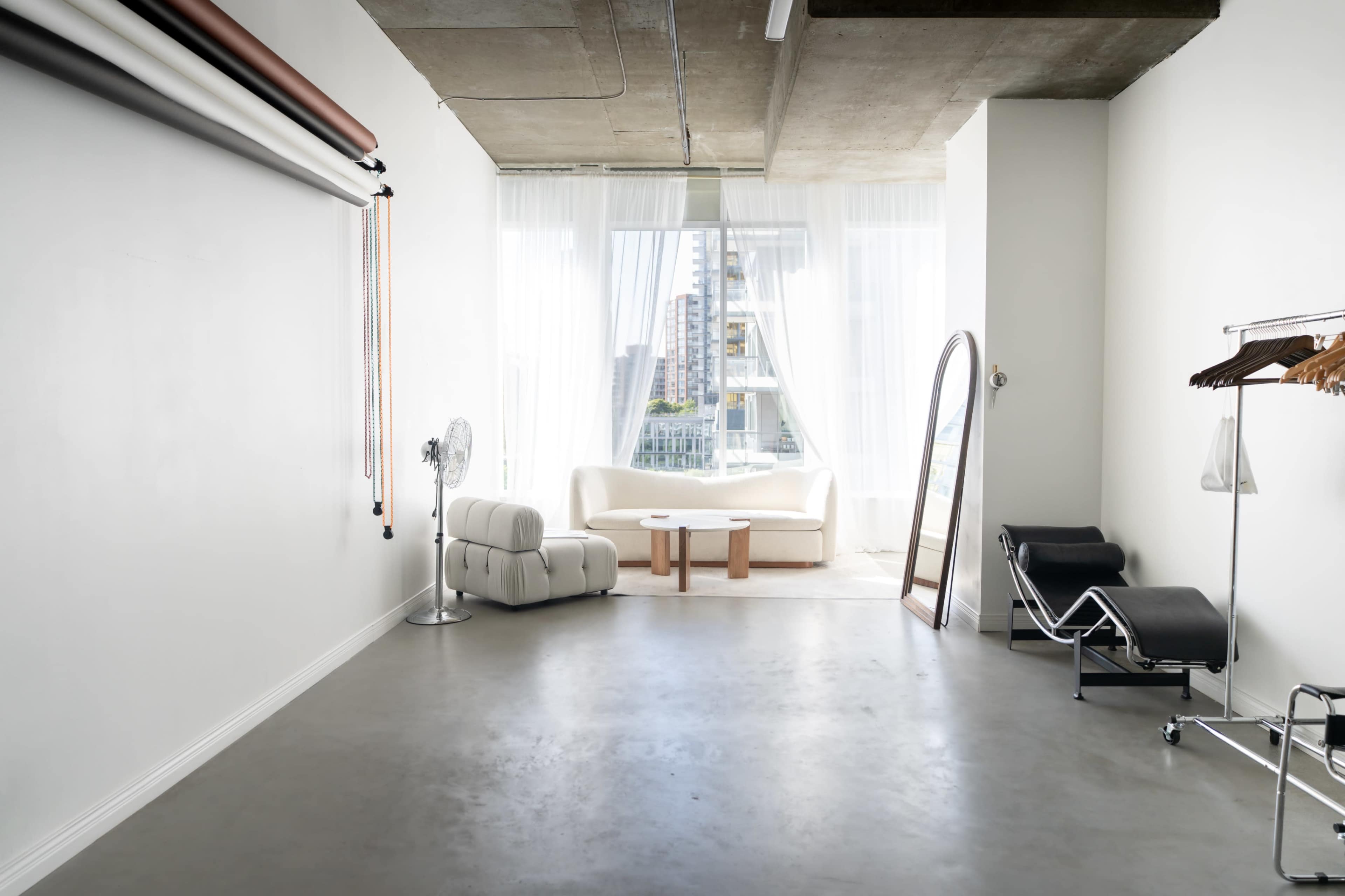 The image shows a minimalist interior space featuring a white couch, large windows with sheer curtains, and a full-length mirror on the right side.