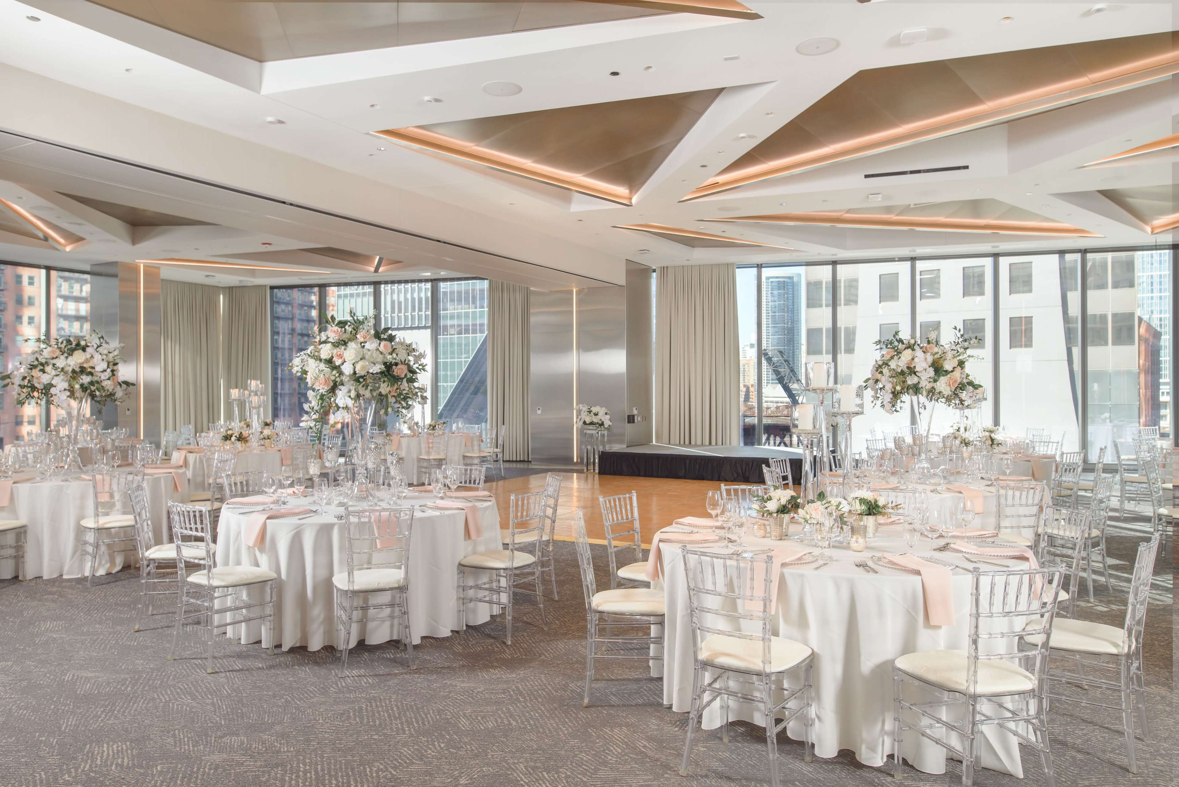 The venue features elegantly arranged round tables with floral centerpieces and a stage, surrounded by large windows showcasing a cityscape.