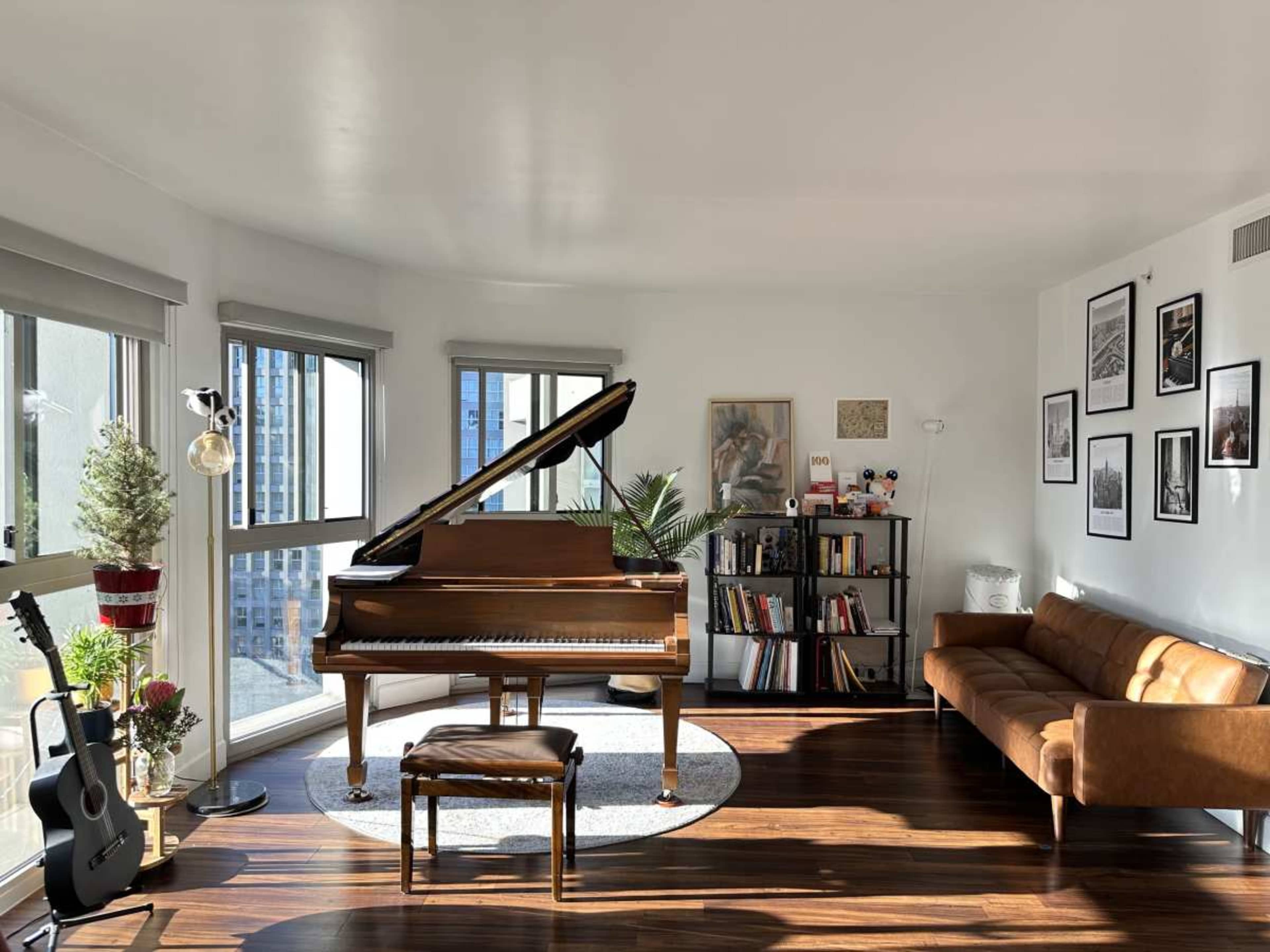 The image shows a well-lit living room featuring a grand piano, a brown leather sofa, a bookshelf, and large windows with city views.