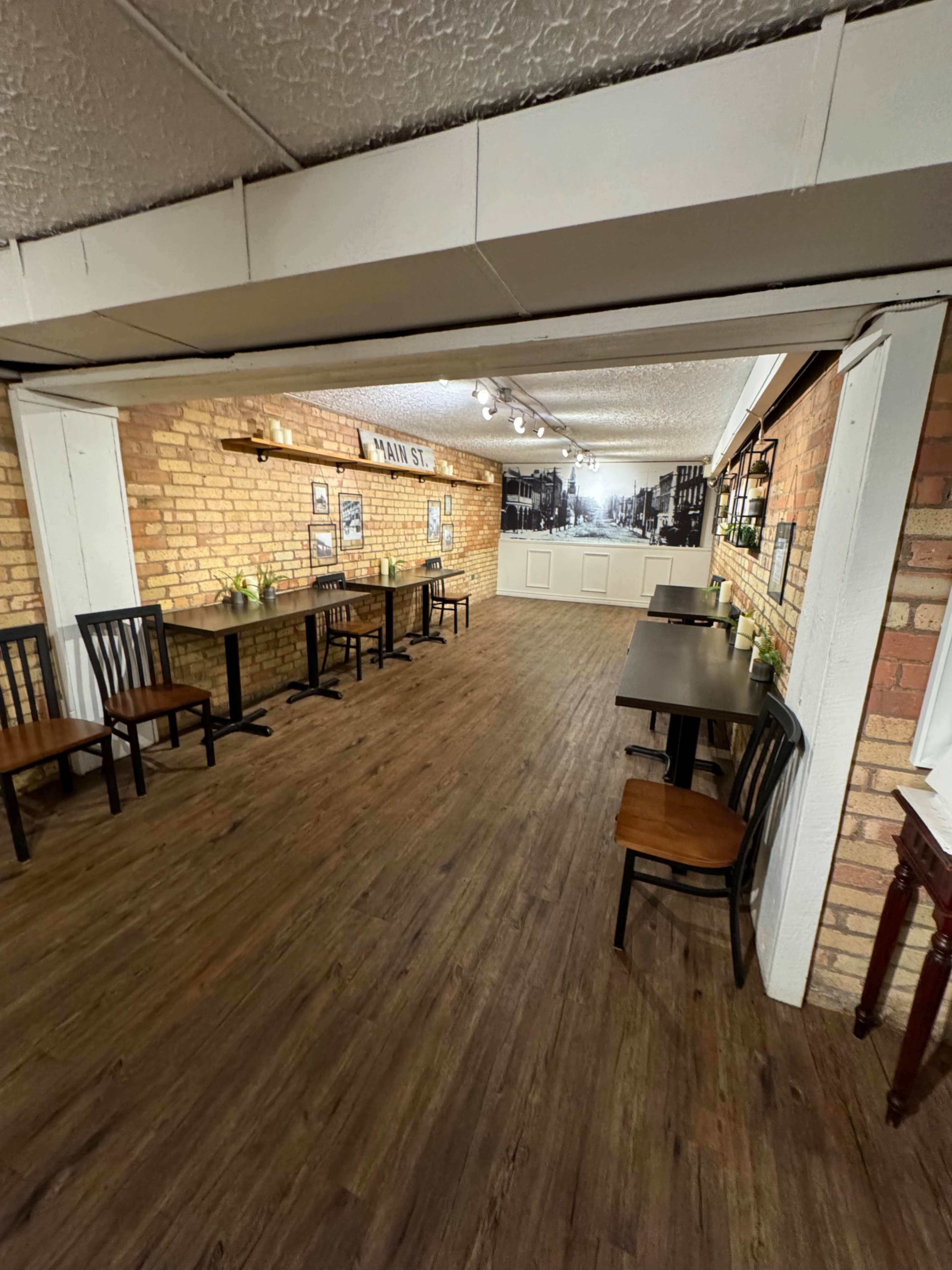 An indoor cafe or restaurant with wooden tables and chairs arranged along a brick wall, featuring framed pictures and a decorative backdrop.