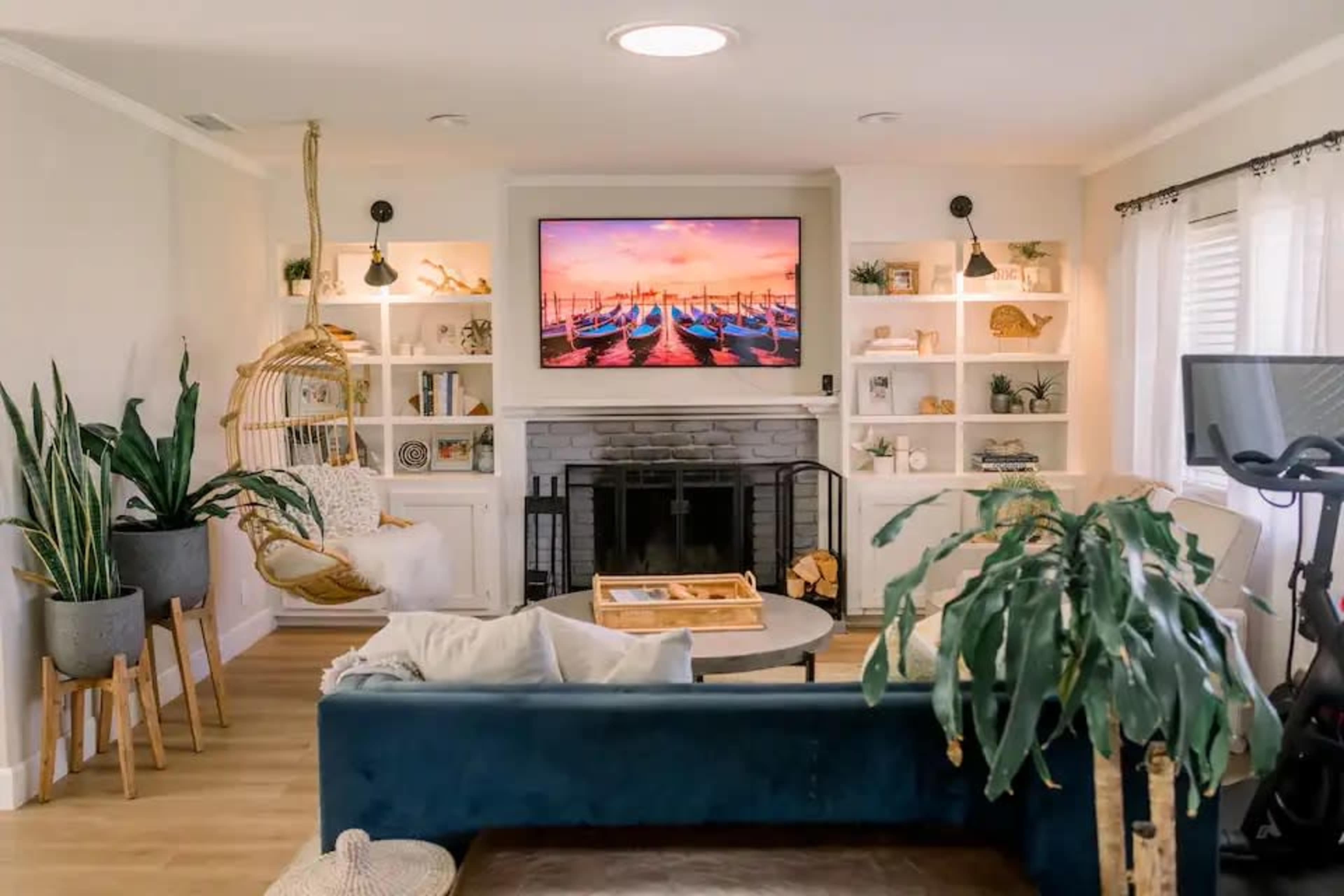 The living room features a navy blue couch, a fireplace with a flat-screen TV mounted above it, shelving with decorative items, and a hanging chair beside a large indoor plant.