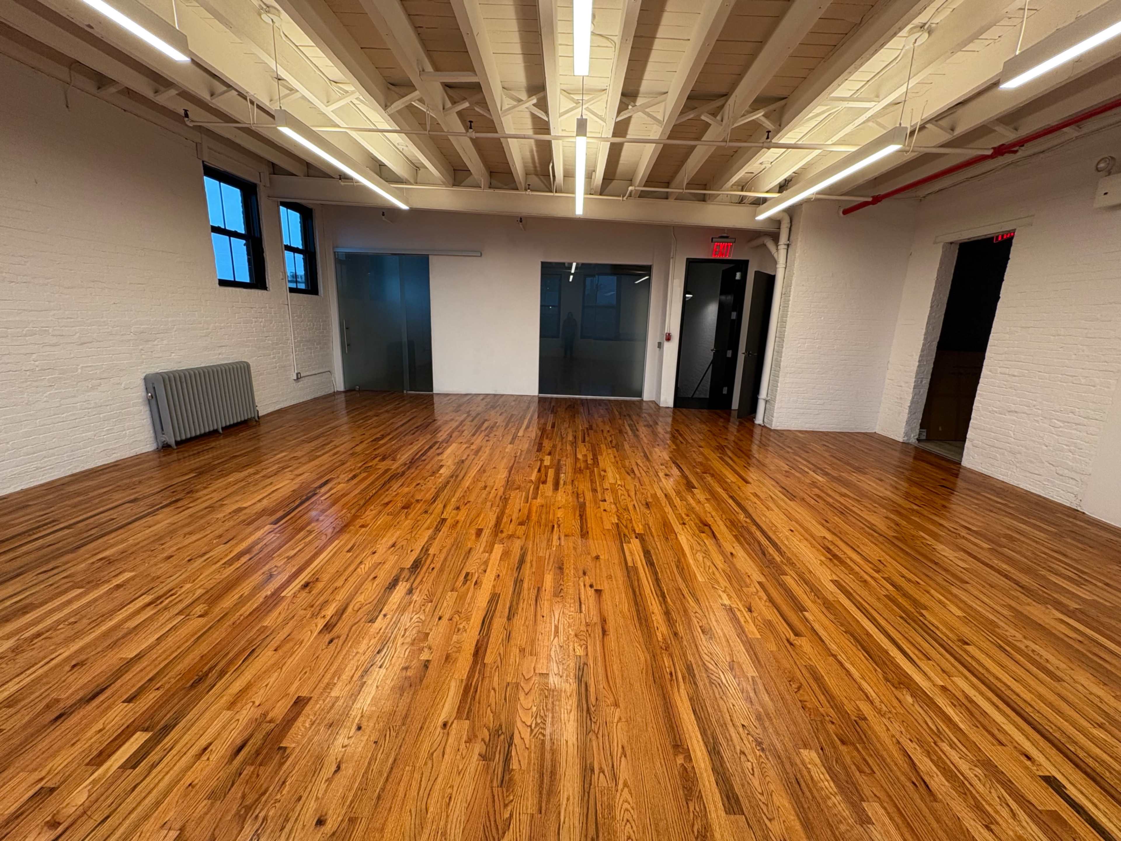 The image shows a large, empty room with polished wooden floors, white brick walls, and multiple windows.