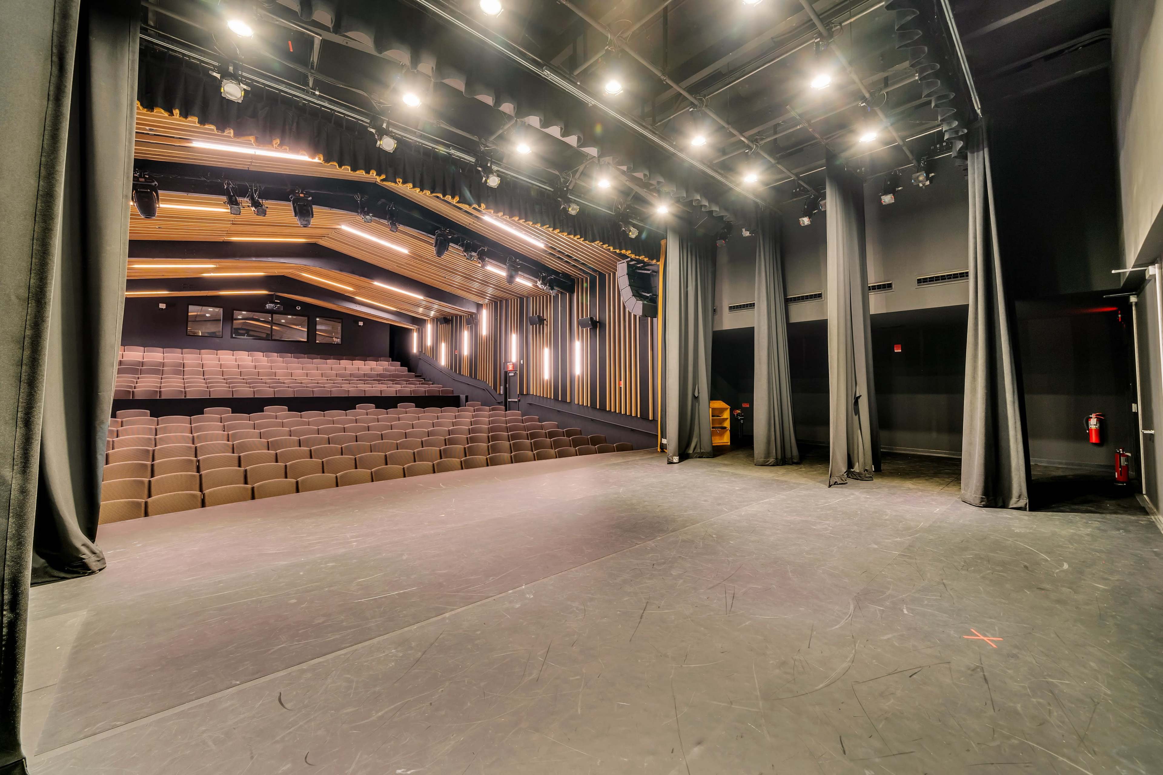 A theater stage with black curtains, illuminated lights, and rows of empty seating in the background.
