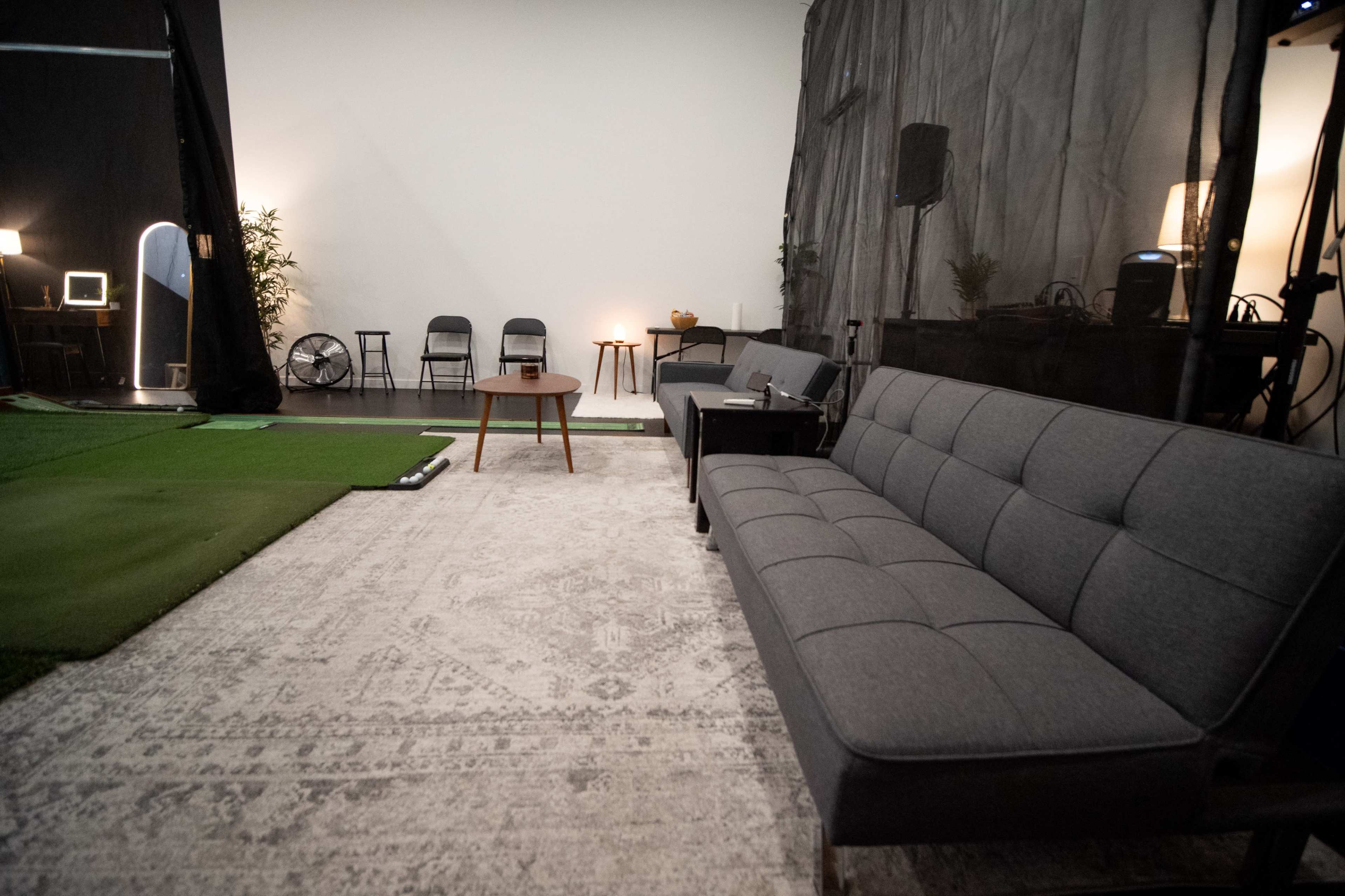 The image shows a modern, minimalistic indoor space featuring a gray sofa, a wooden coffee table, and a small area of green turf on the floor.