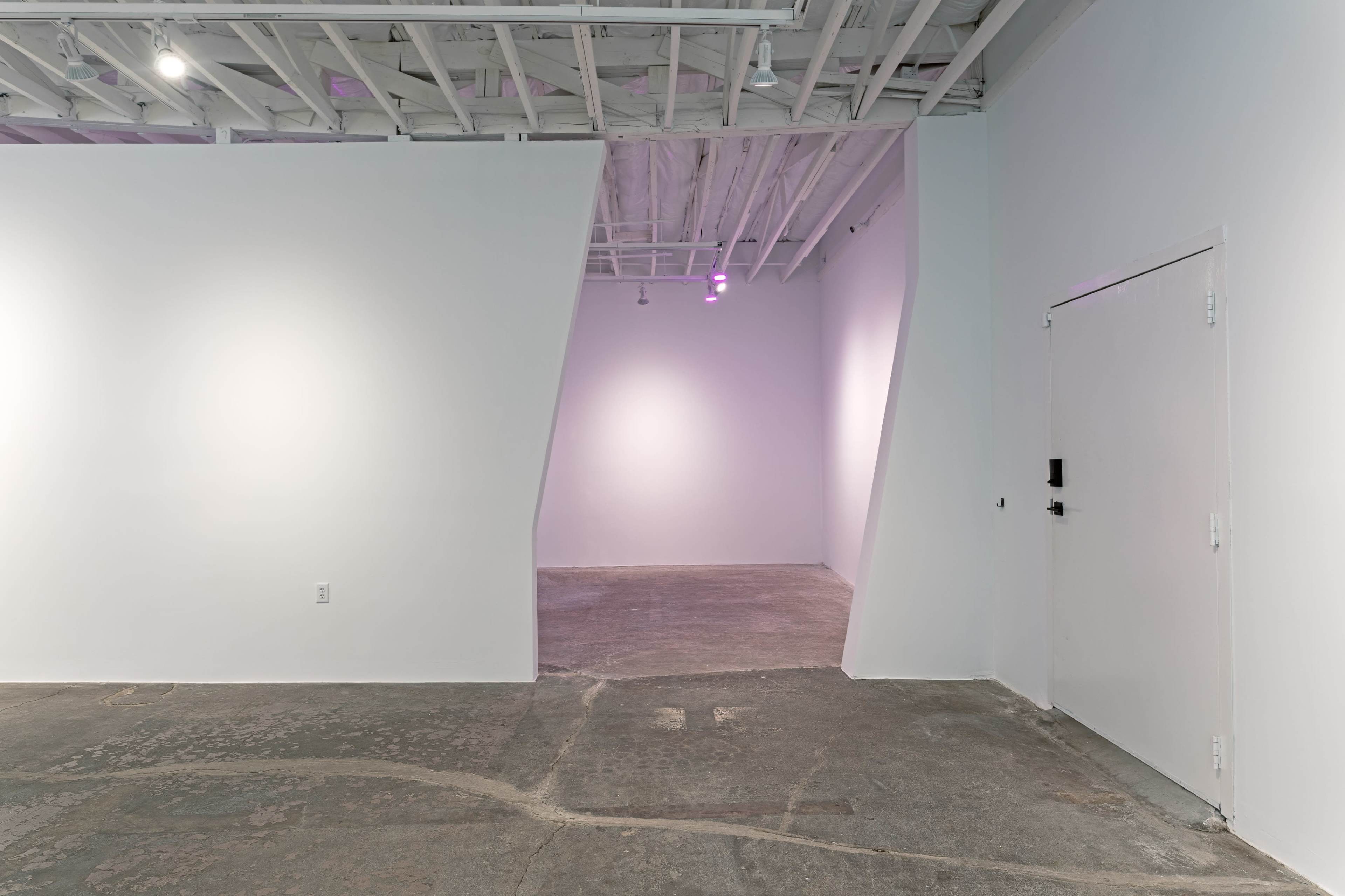 A minimalist interior space with white walls, a concrete floor, and an angled partition leading to another room.