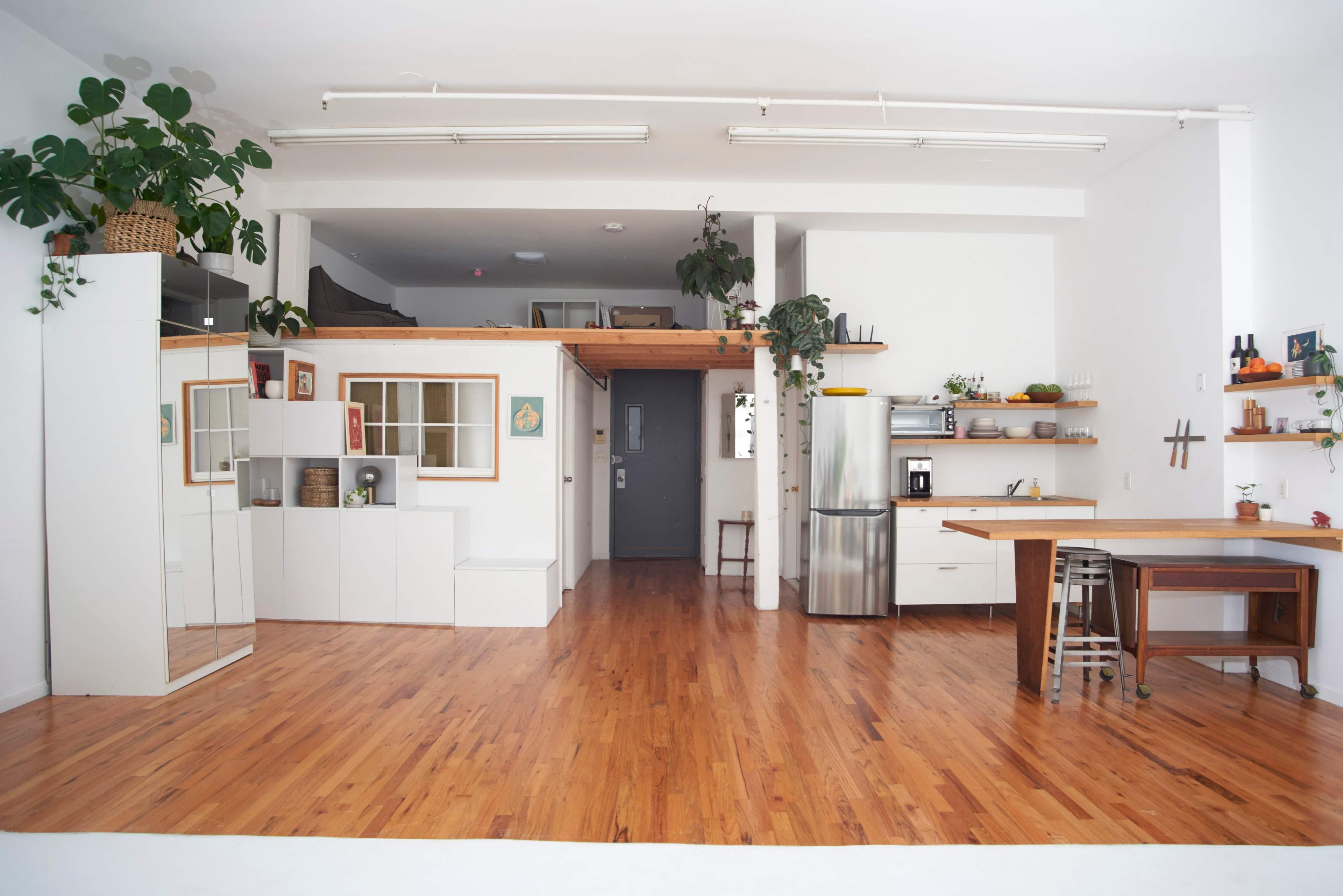The image shows a large, open-concept space with wooden flooring, a kitchen area, and a lofted section adorned with plants.