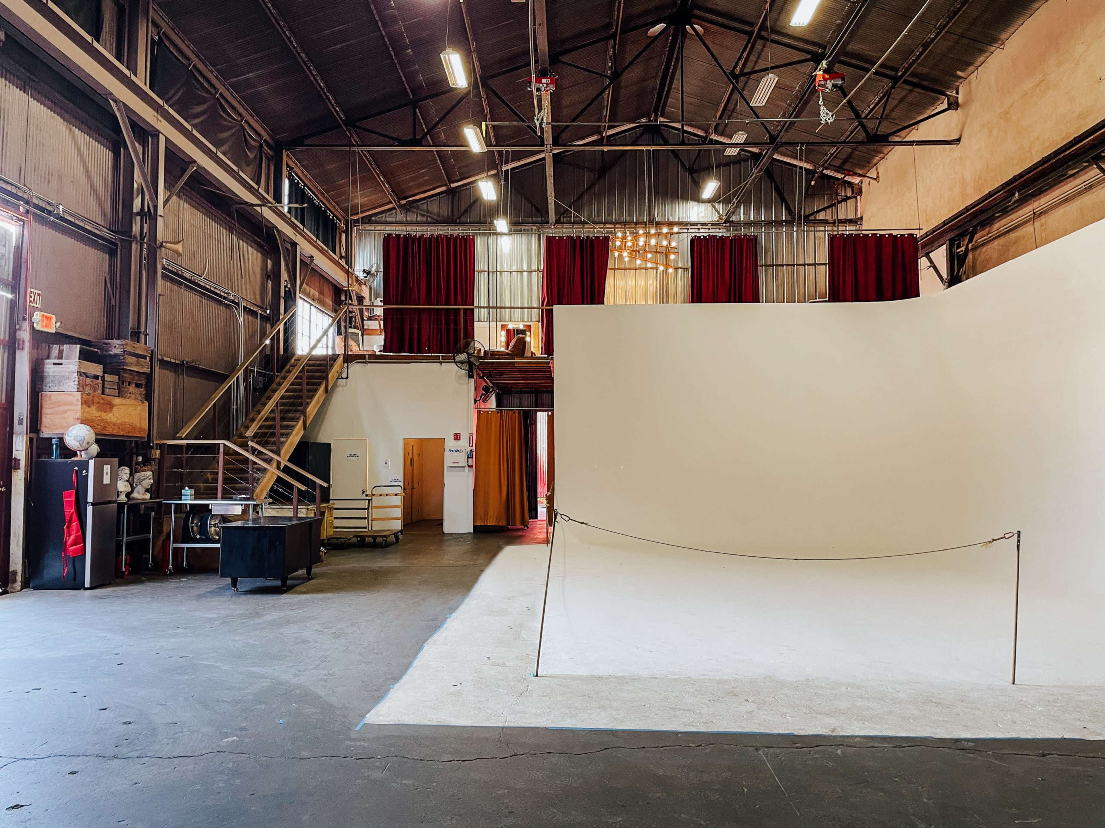 The image shows a spacious industrial studio with a large empty white backdrop, red curtains, and a staircase leading to an upper level.