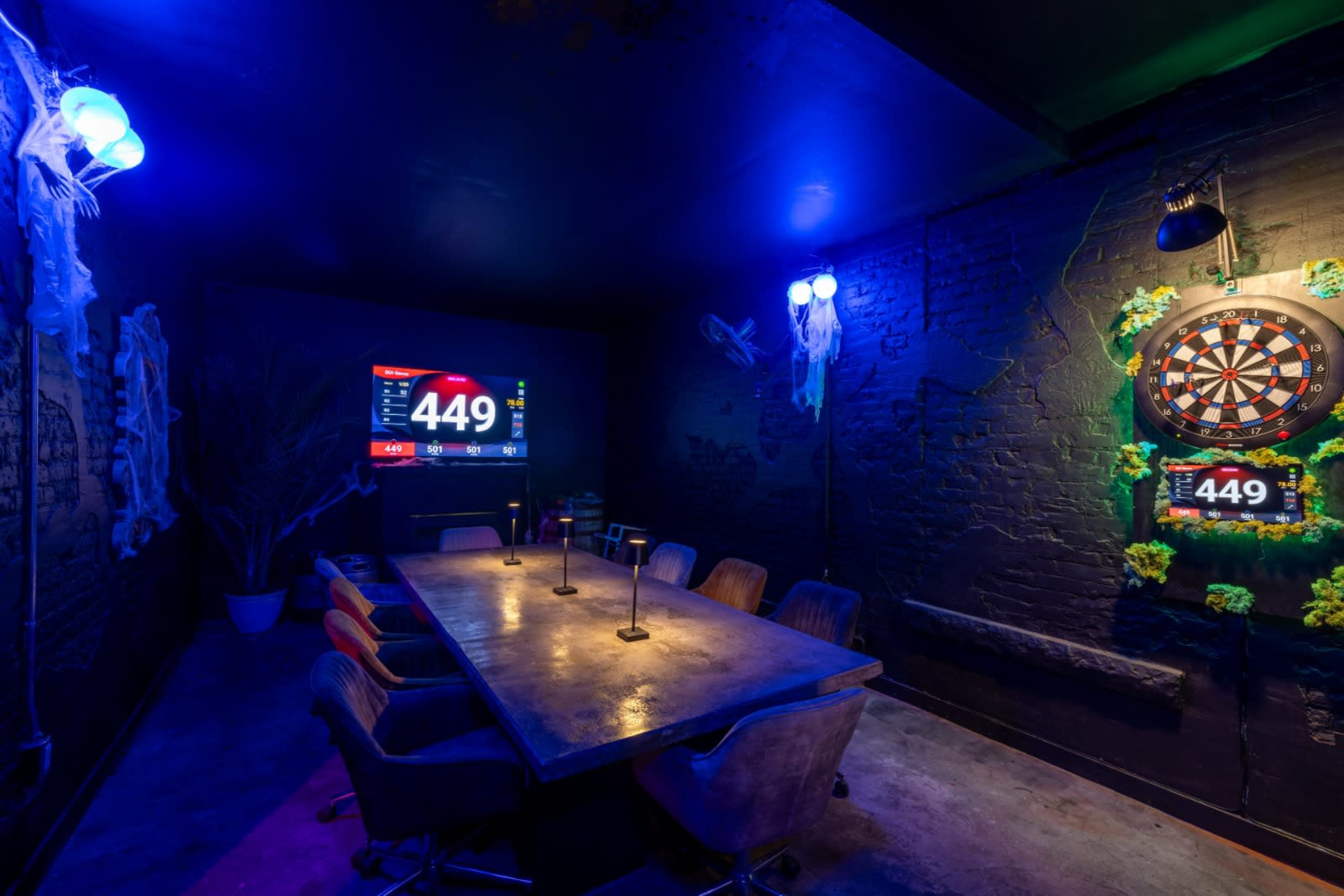A dimly lit room features a large table surrounded by chairs, with a dartboard and a screen displaying a score.