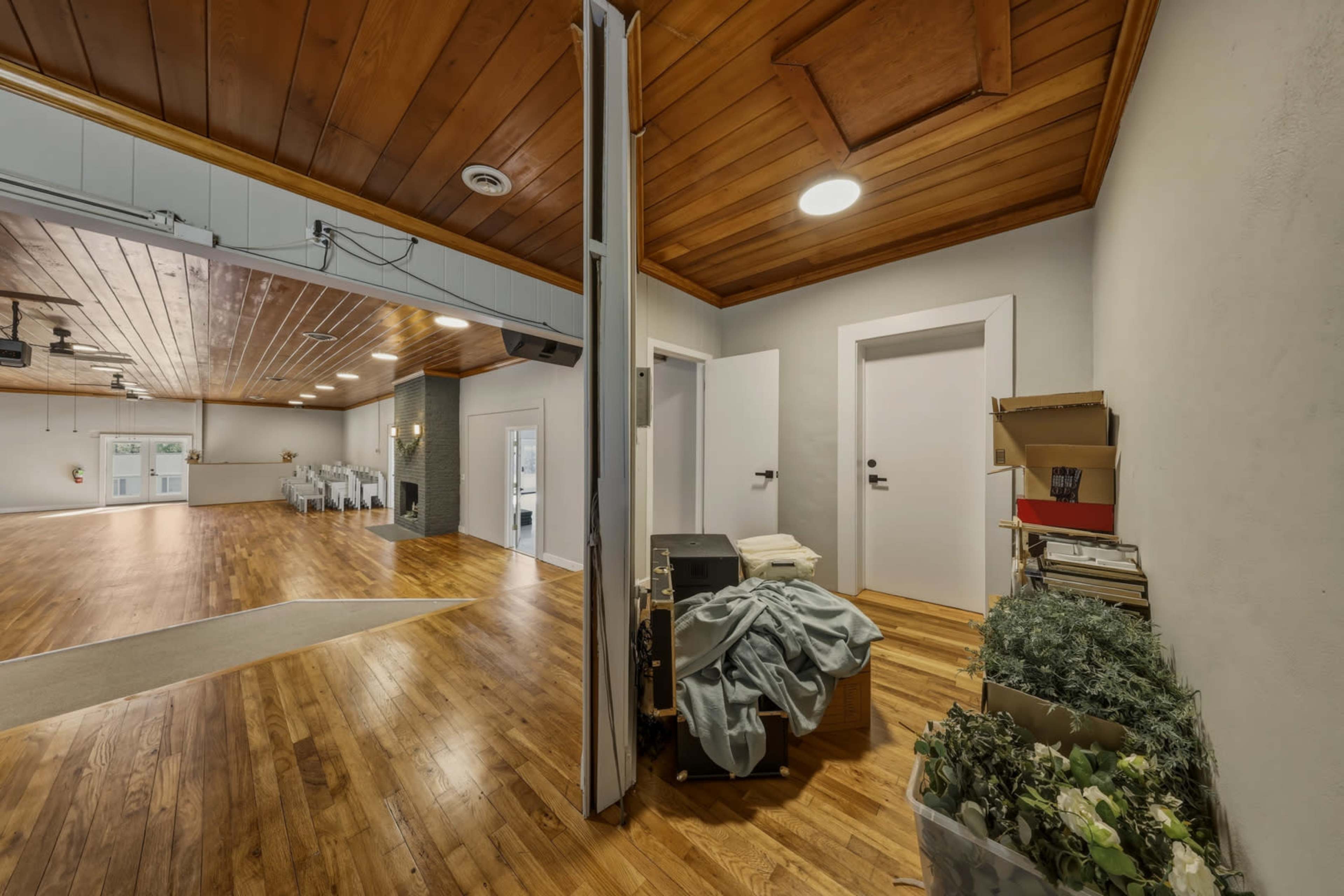 The image shows a spacious interior with wooden flooring, featuring an entrance to a large room and a storage area containing boxes and plants along one wall.
