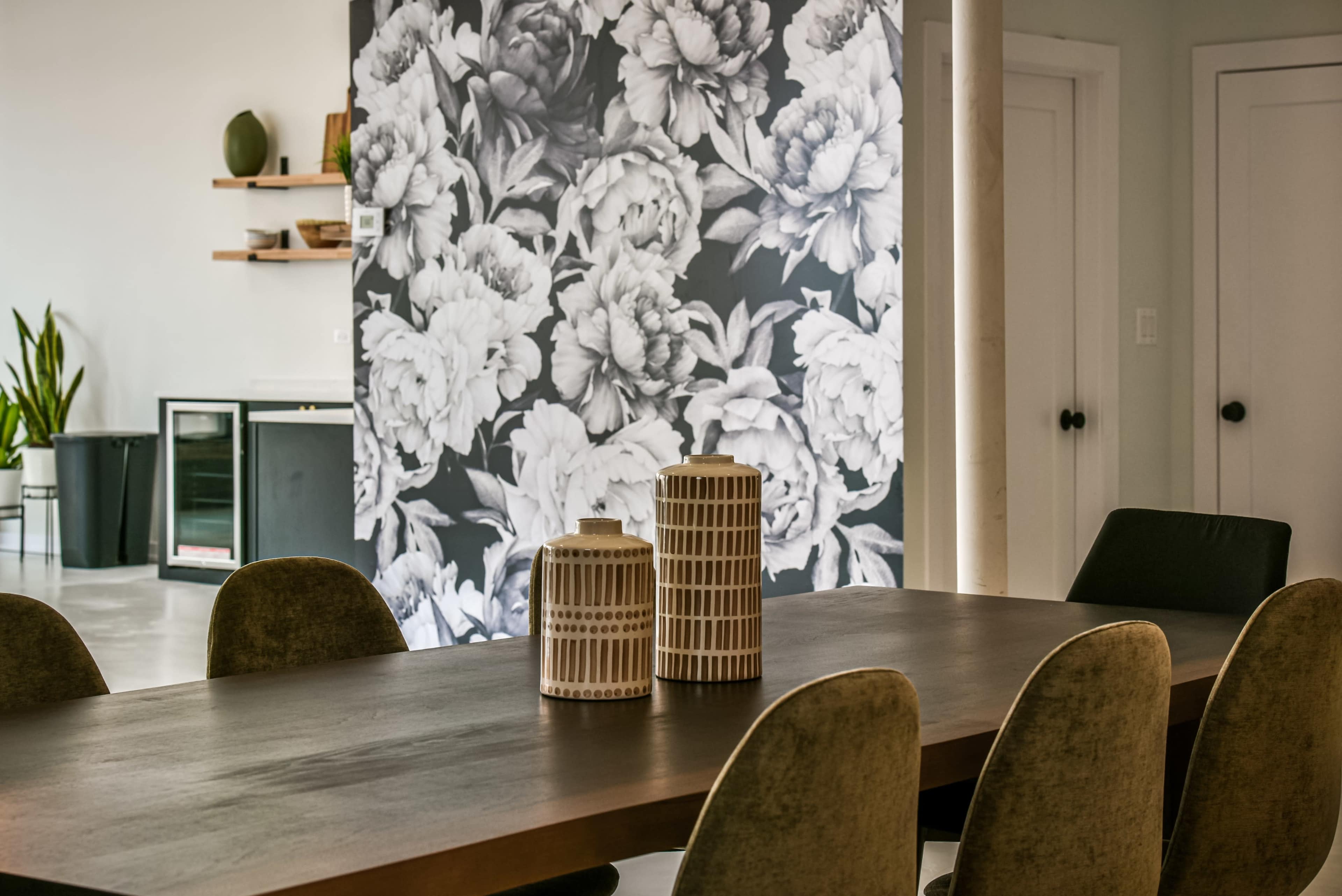 A dining table with decorative vases is set in a room featuring a floral wallpaper and minimalist decor.