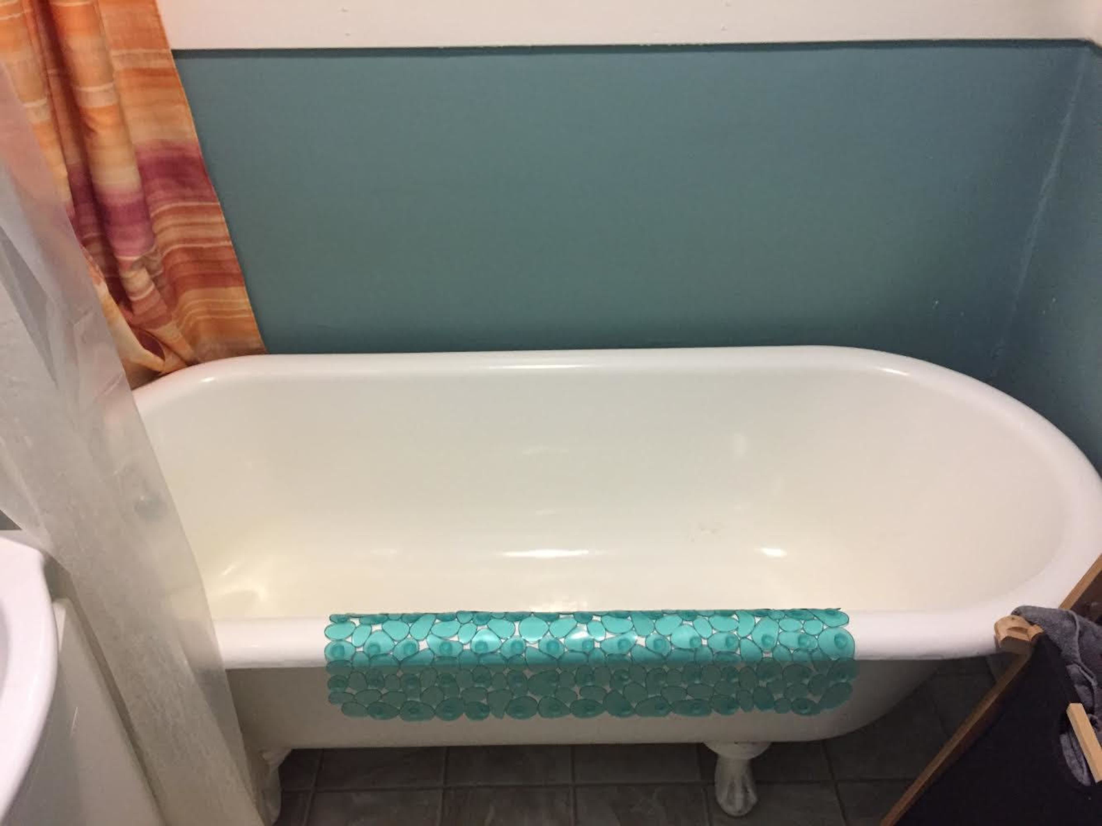 The image shows a white bathtub with decorative blue tiles placed along the edge, situated in a bathroom with a colorful curtain and turquoise wall.