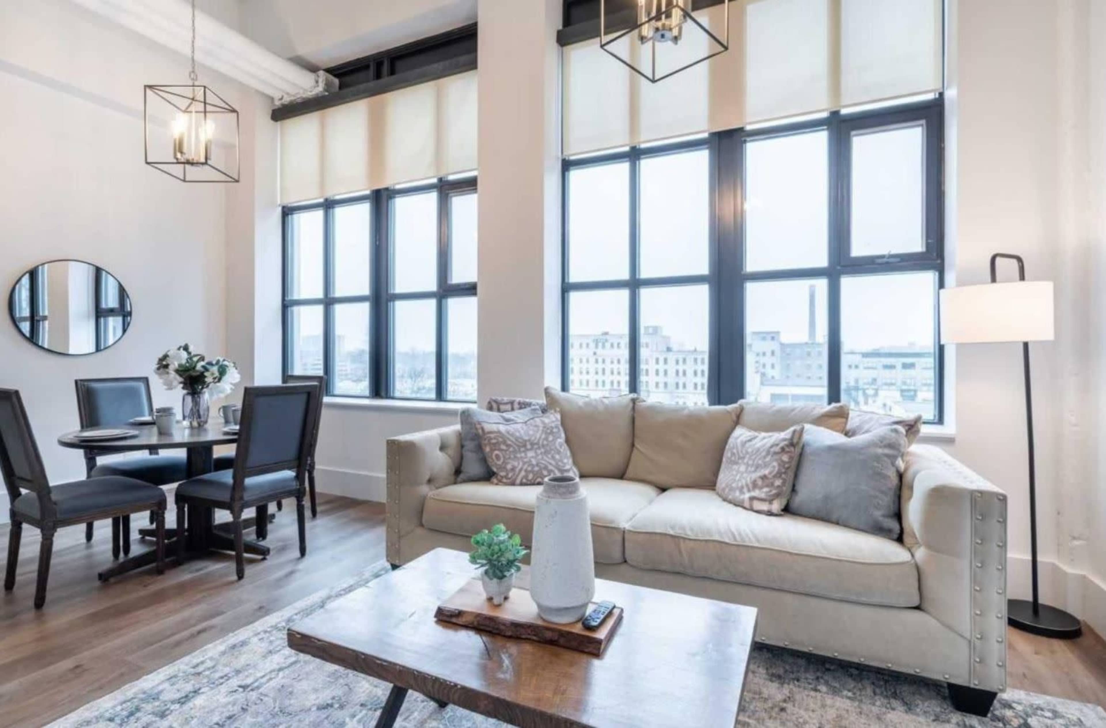 A modern living area featuring a beige sofa, a wooden coffee table, a round dining table with chairs, and large windows allowing natural light to fill the space.