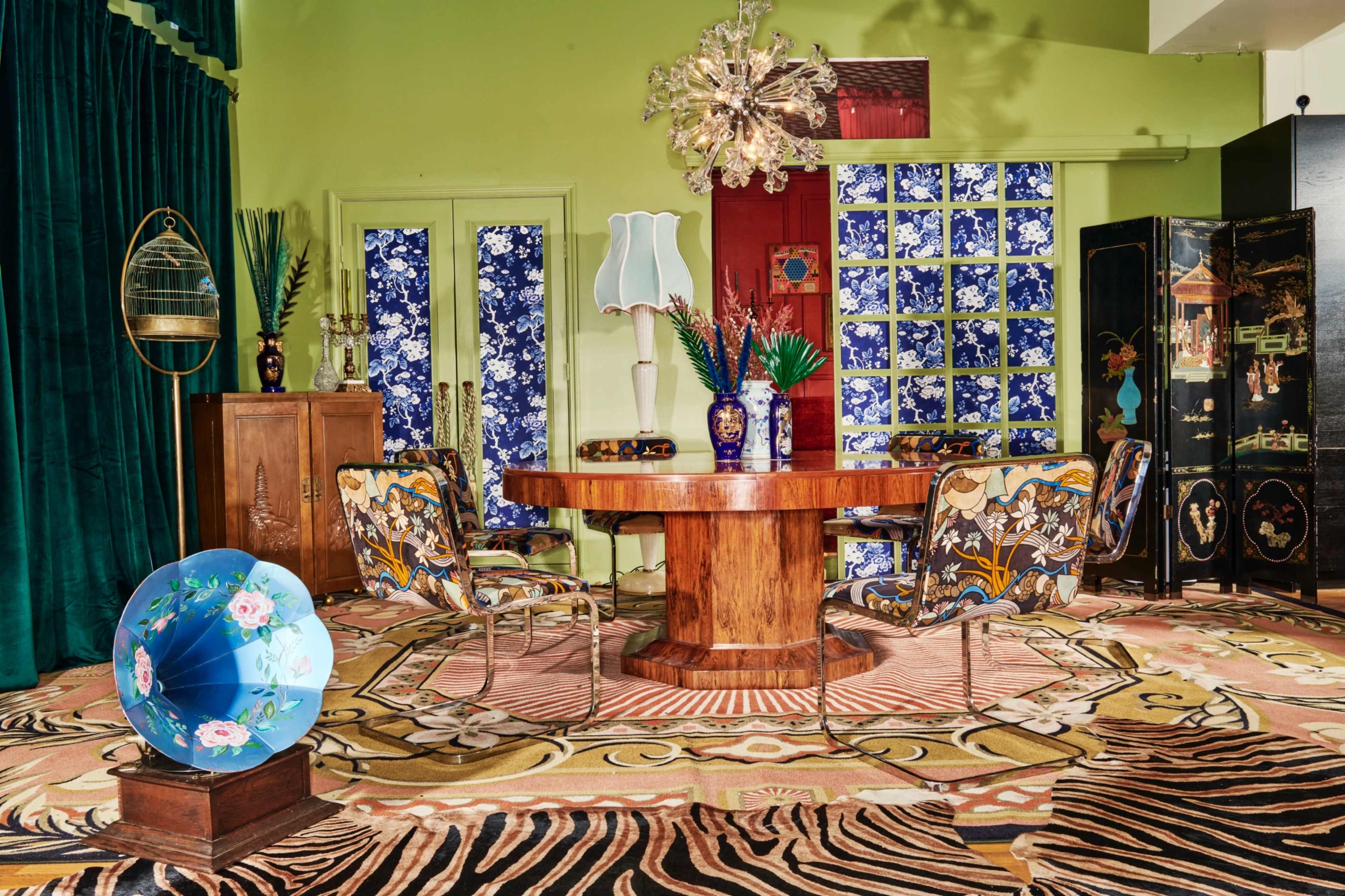 A dining area features a round wooden table surrounded by patterned chairs, with vibrant wallpaper and decorative accessories including a globe and a chandelier.