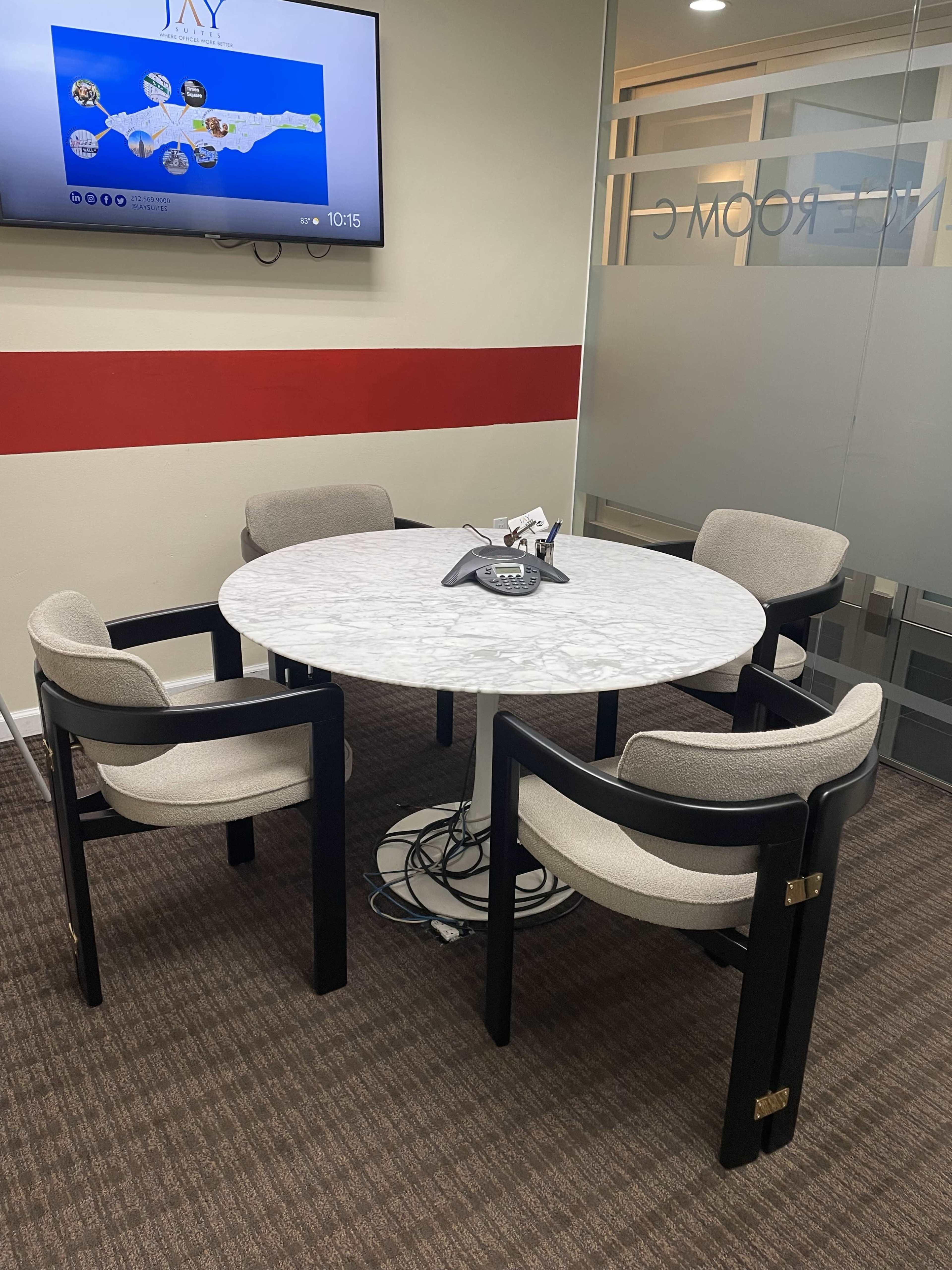A round marble table with four cushioned chairs is positioned in a conference room with a television displaying a map.