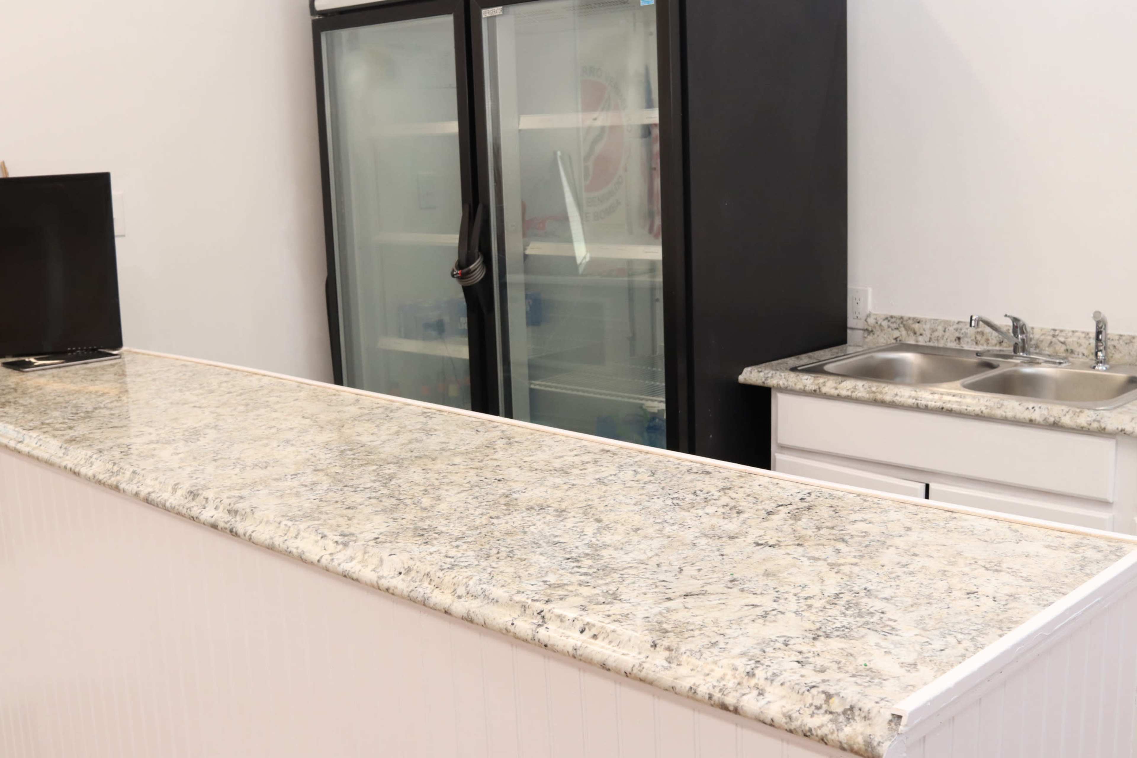 A countertop with a speckled granite surface is positioned in front of a black refrigerator and a double sink against a white wall.
