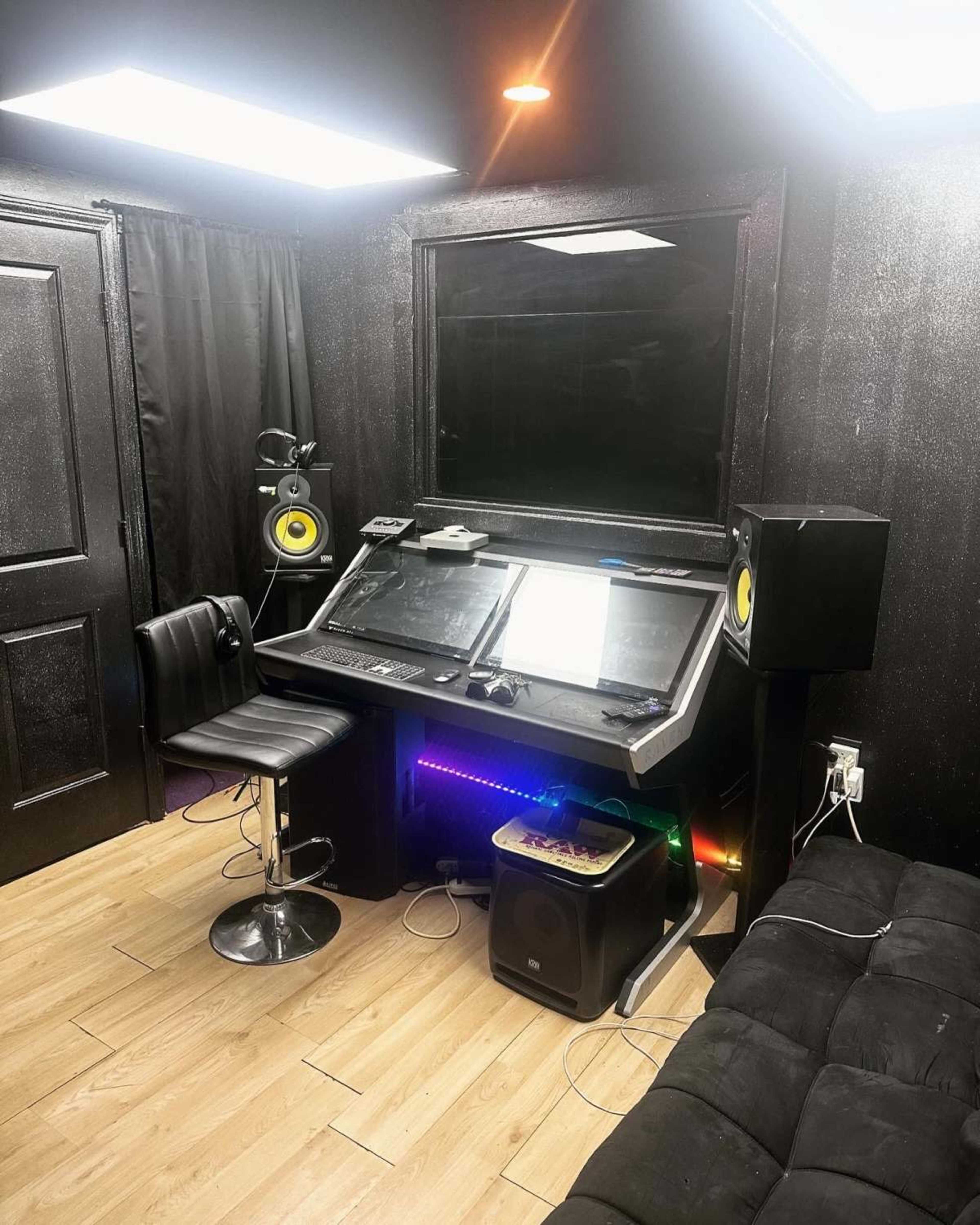 The image shows a modern music production workspace featuring a sleek desk with monitors, studio speakers, and a black swivel chair in a dimly lit room with wooden flooring.