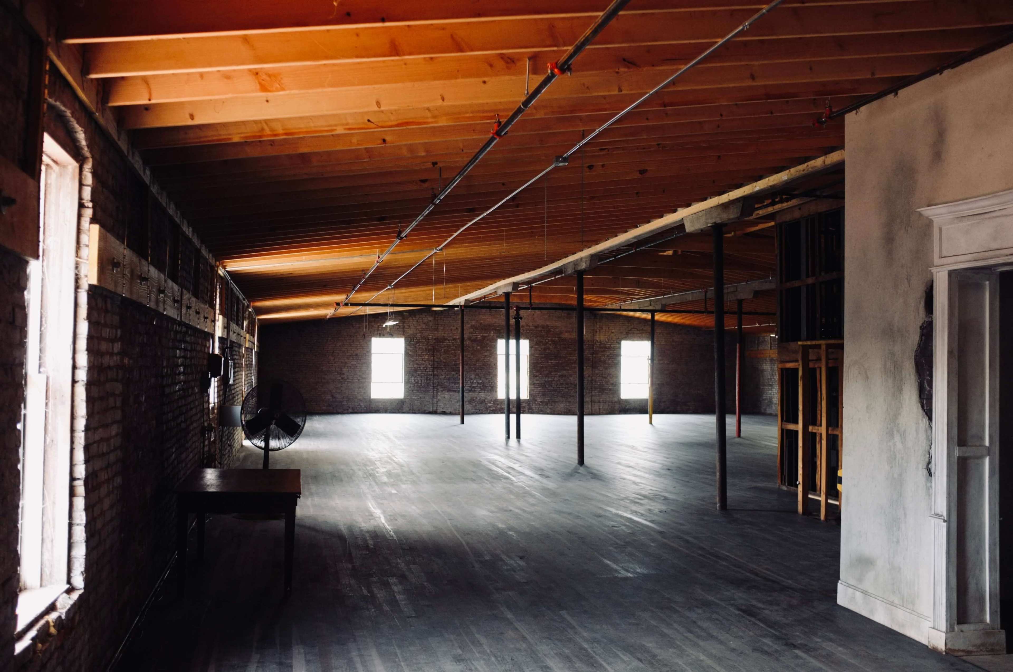 An empty industrial space features exposed brick walls, wooden beams, and large windows allowing natural light to enter.