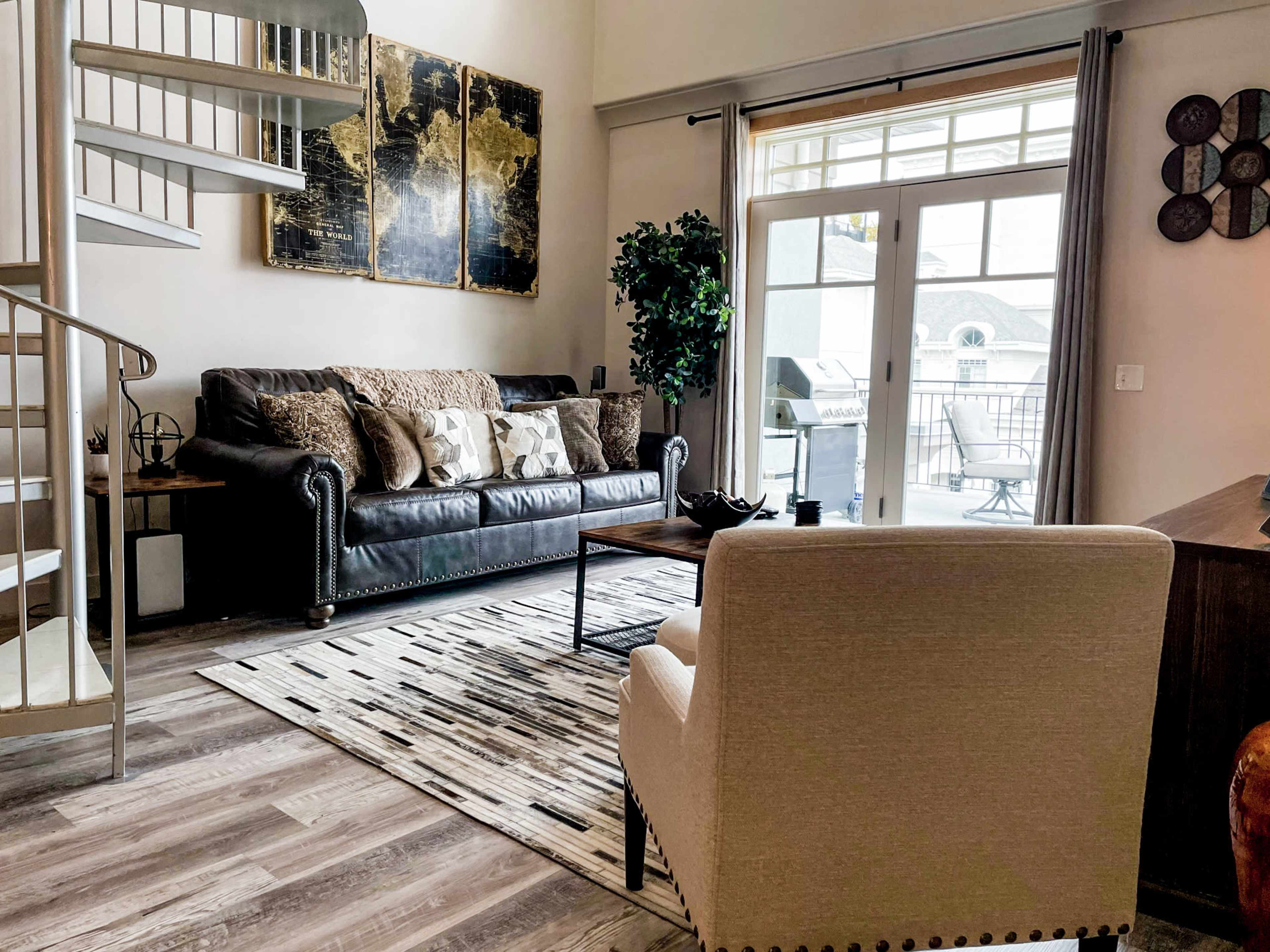 A modern living room with a spiral staircase, a black leather couch adorned with decorative pillows, and a balcony visible through large glass doors.