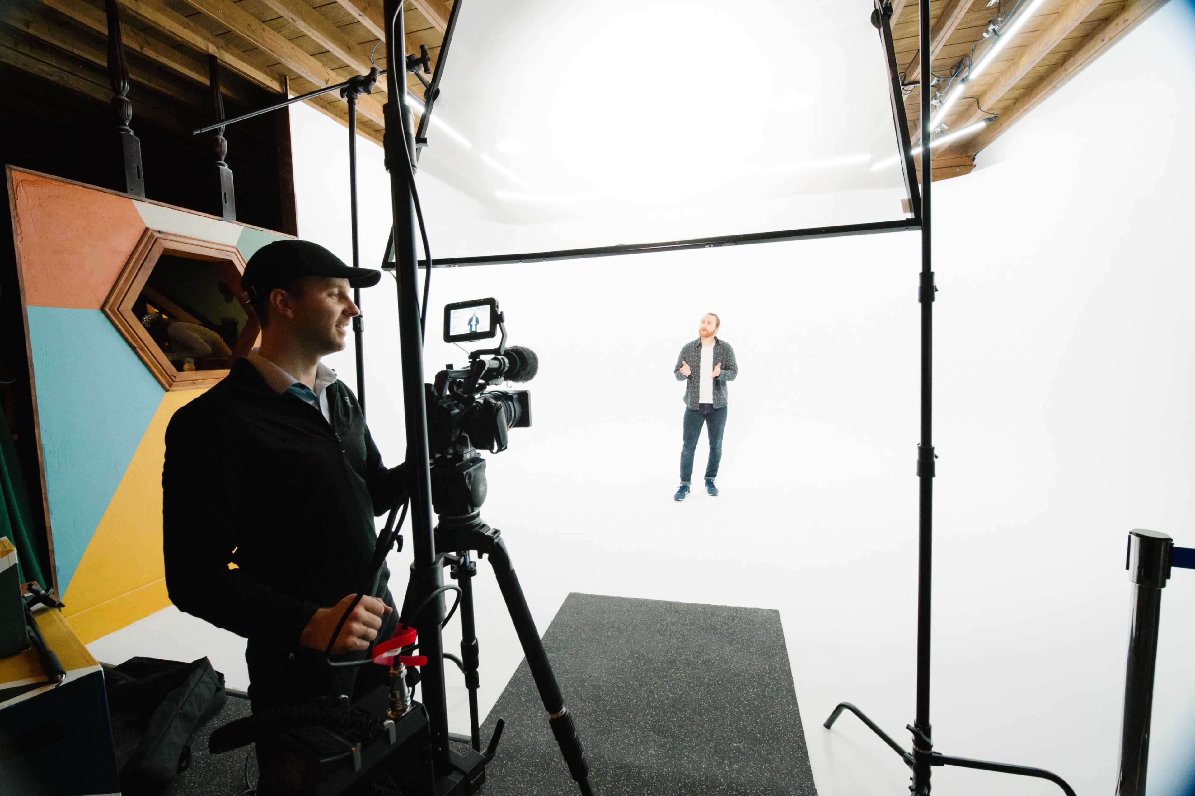 A man stands on stage in front of a white backdrop while another man operates a camera on a tripod.