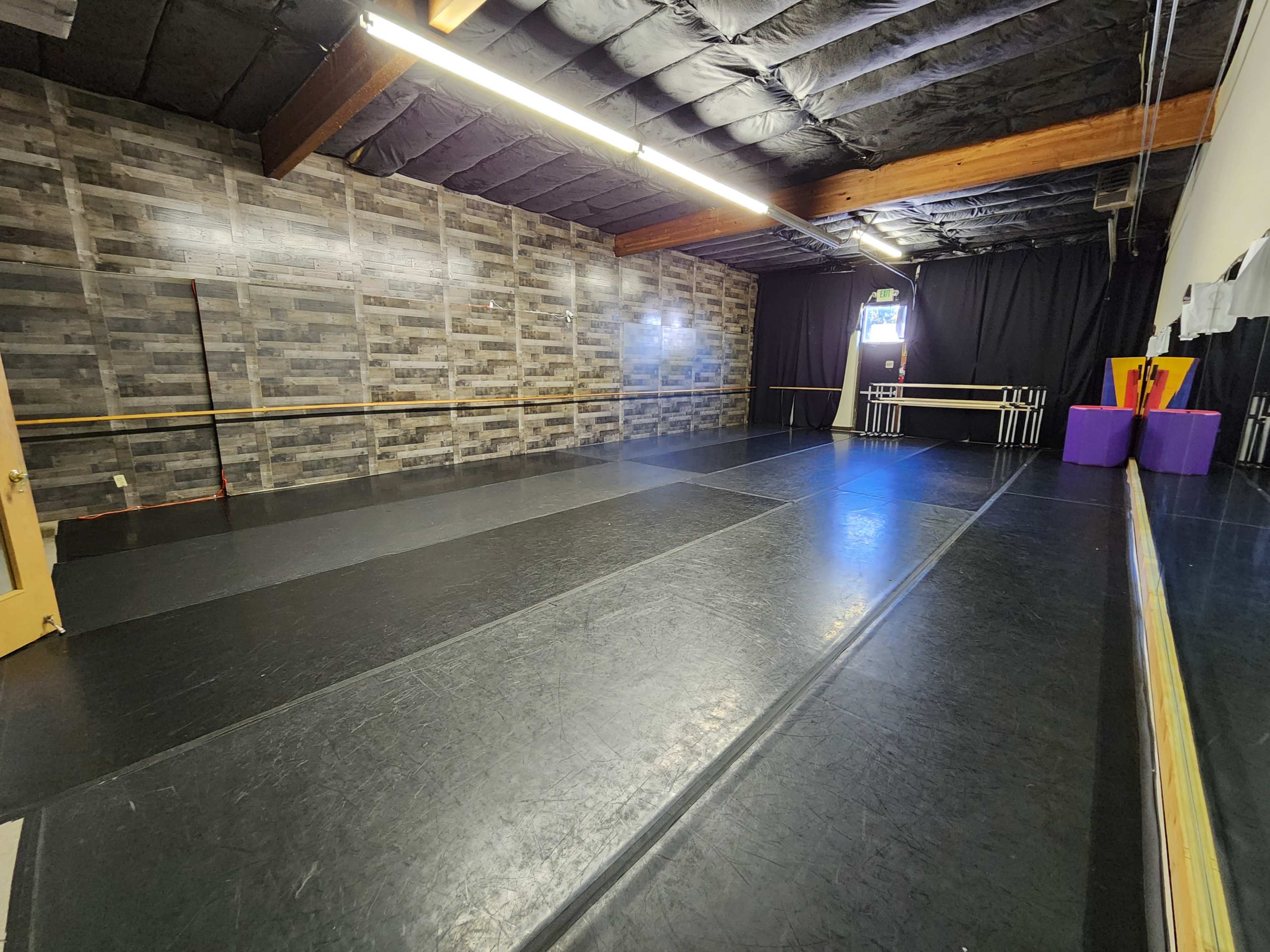 The image shows an empty dance studio with a black floor, mirrored wall, and a wooden ceiling, along with a textured wall in the background.