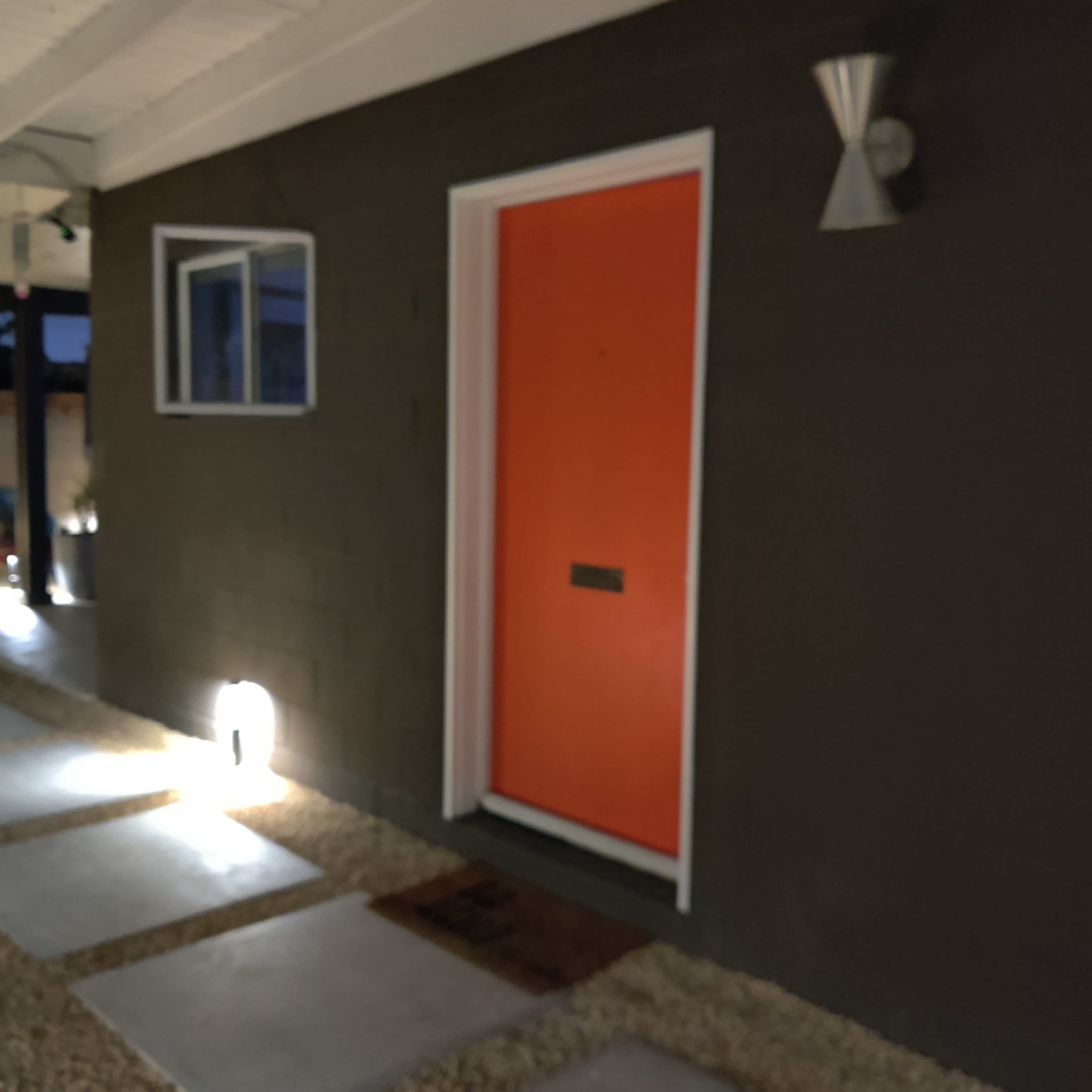 The image shows an orange door with a mail slot, situated on a dark gray exterior wall, accompanied by a wall-mounted light fixture beside a small window.