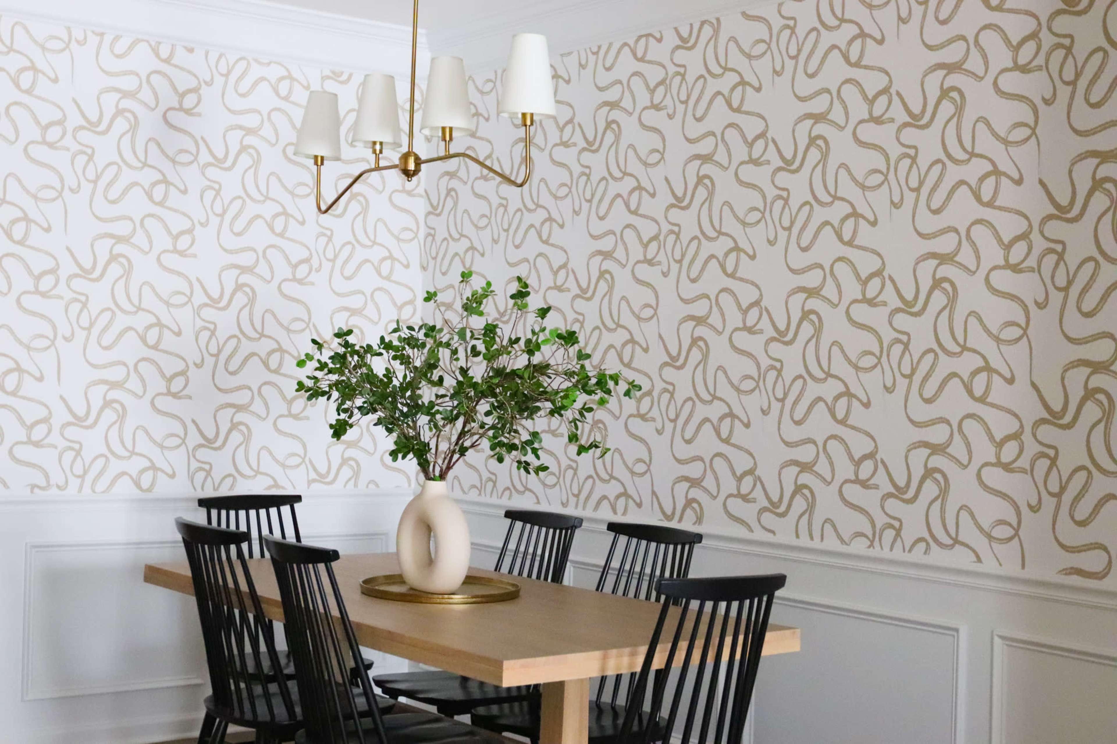 A dining area features a wooden table surrounded by black chairs, topped with a decorative vase and a green plant, against a patterned wallpaper backdrop.