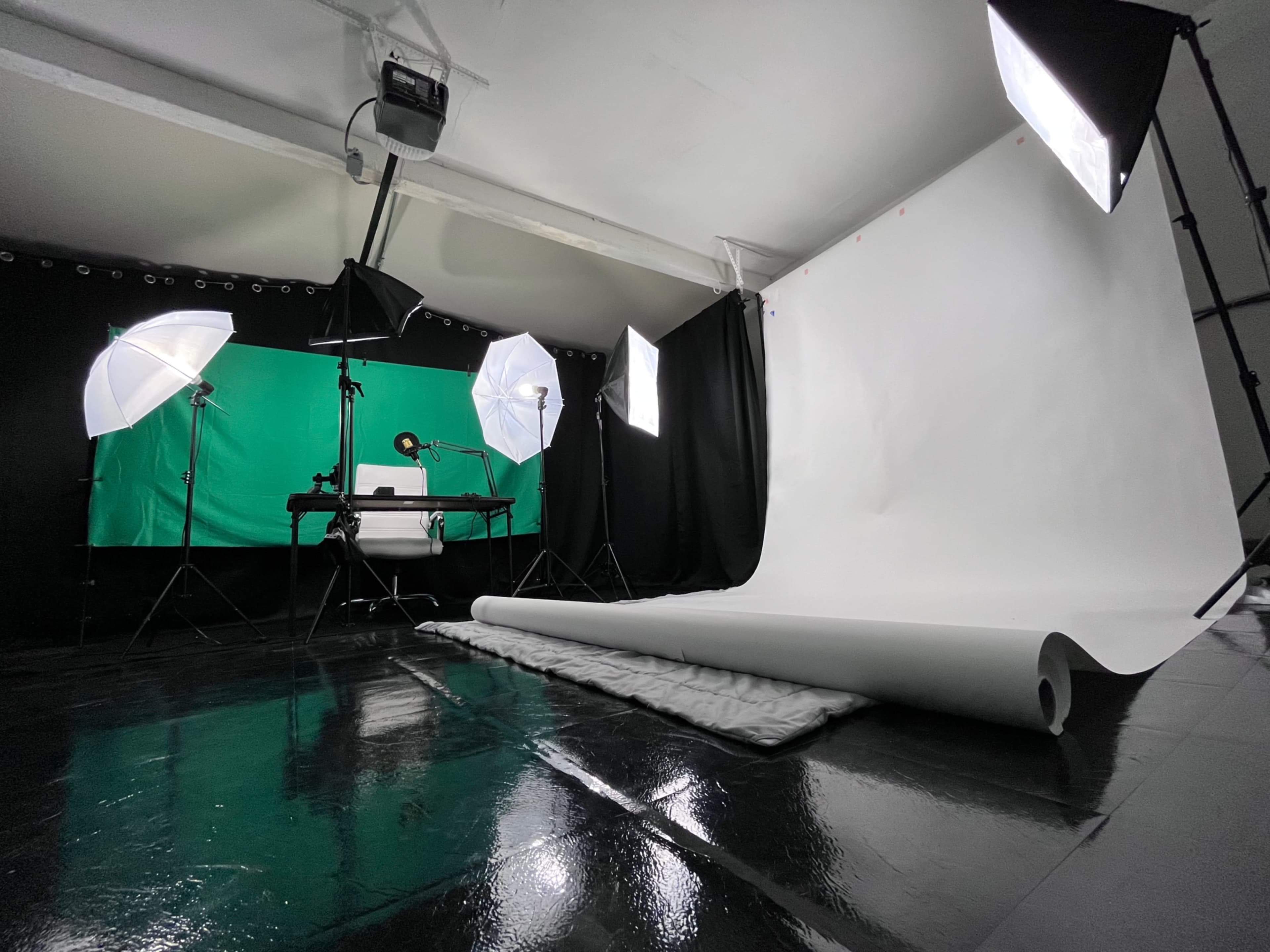 A photography studio setup with a white backdrop, green screen, lighting equipment, and a table for photo shoots.