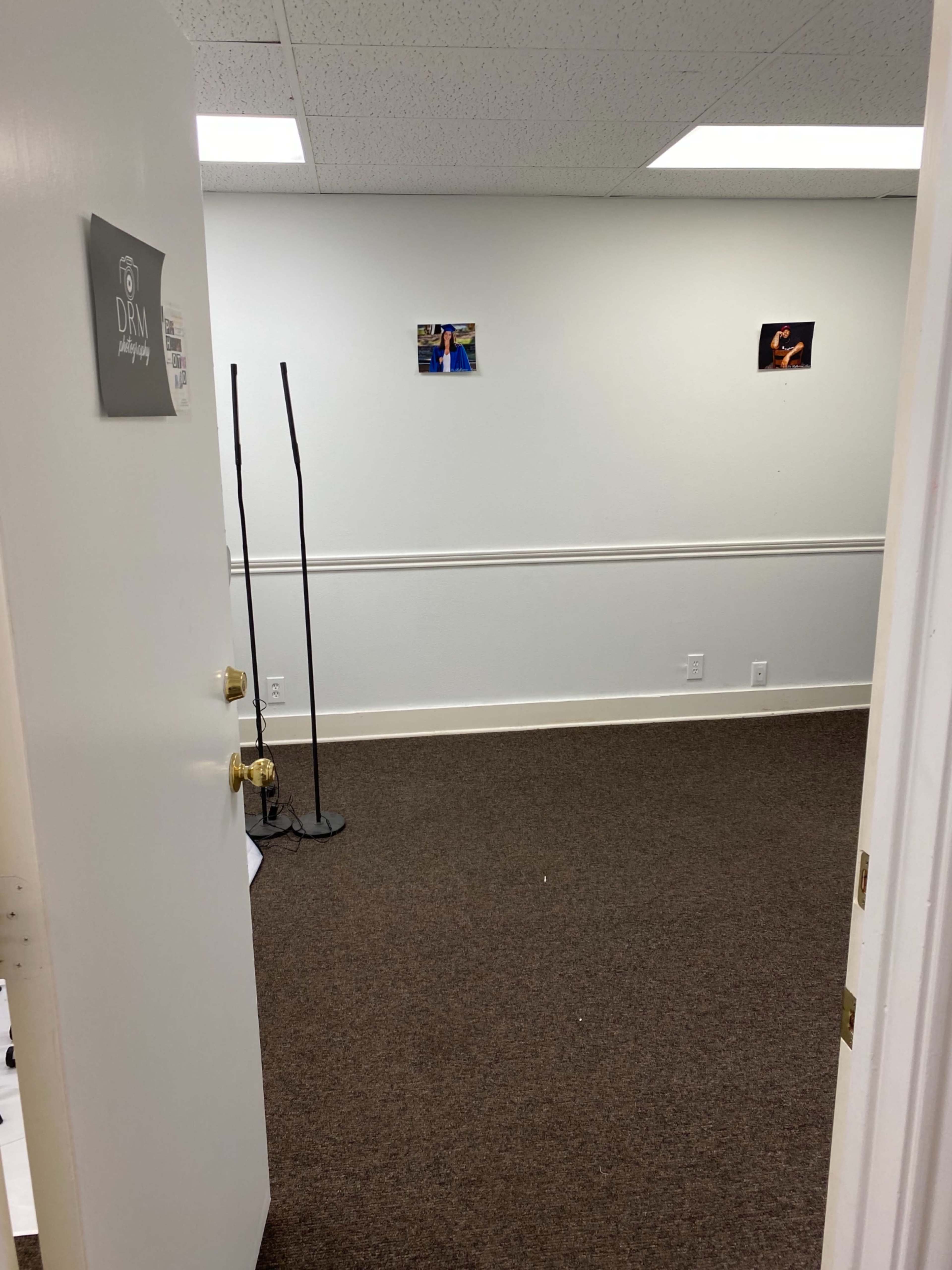 An empty office space with beige carpet, white walls featuring a couple of photographs, and a door partially open.