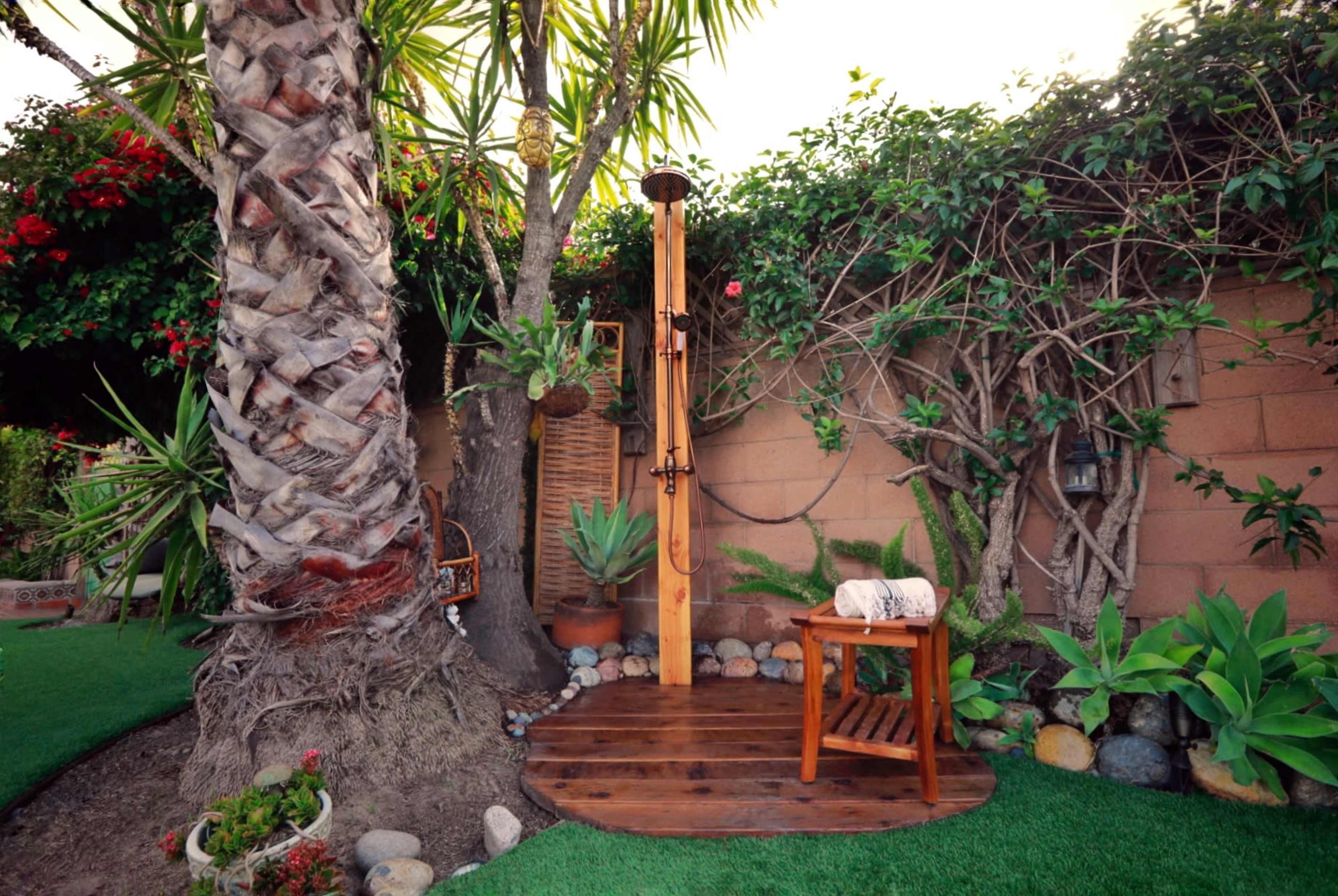 A wooden outdoor shower is set on a circular wooden platform surrounded by tropical plants and a palm tree.