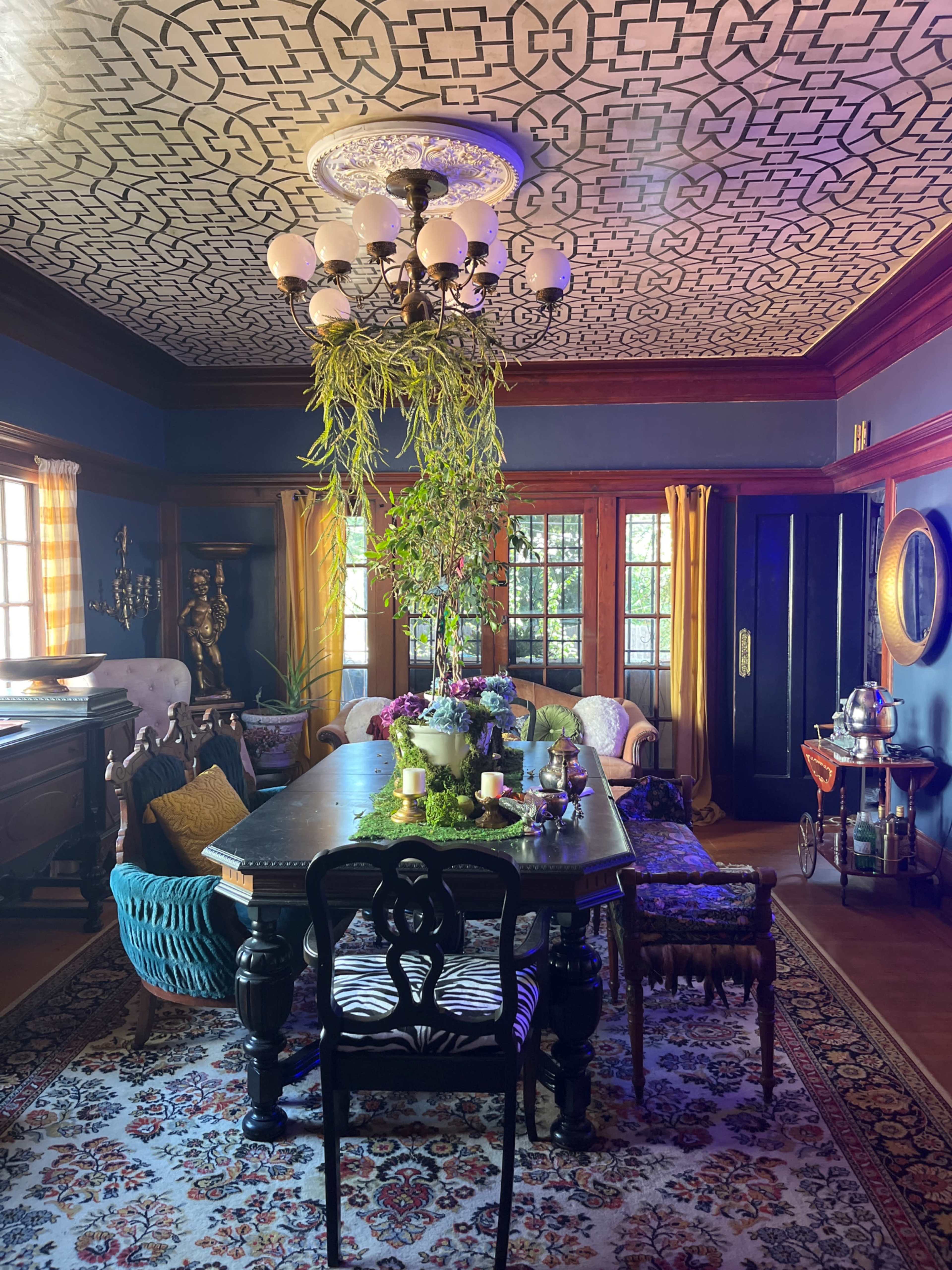 The image shows a richly decorated dining room with a patterned ceiling, a large table at the center, and various plants and furniture arranged around the space.