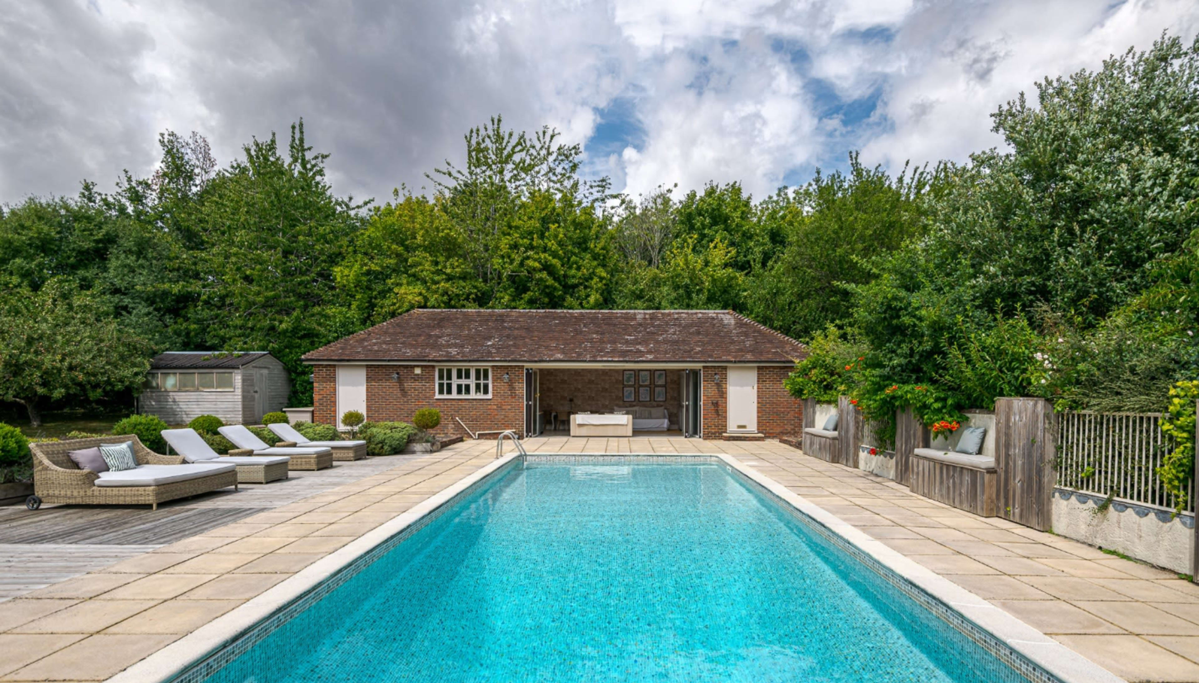 A rectangular swimming pool is surrounded by lounge chairs and a brick building set amidst lush green trees under a cloudy sky.