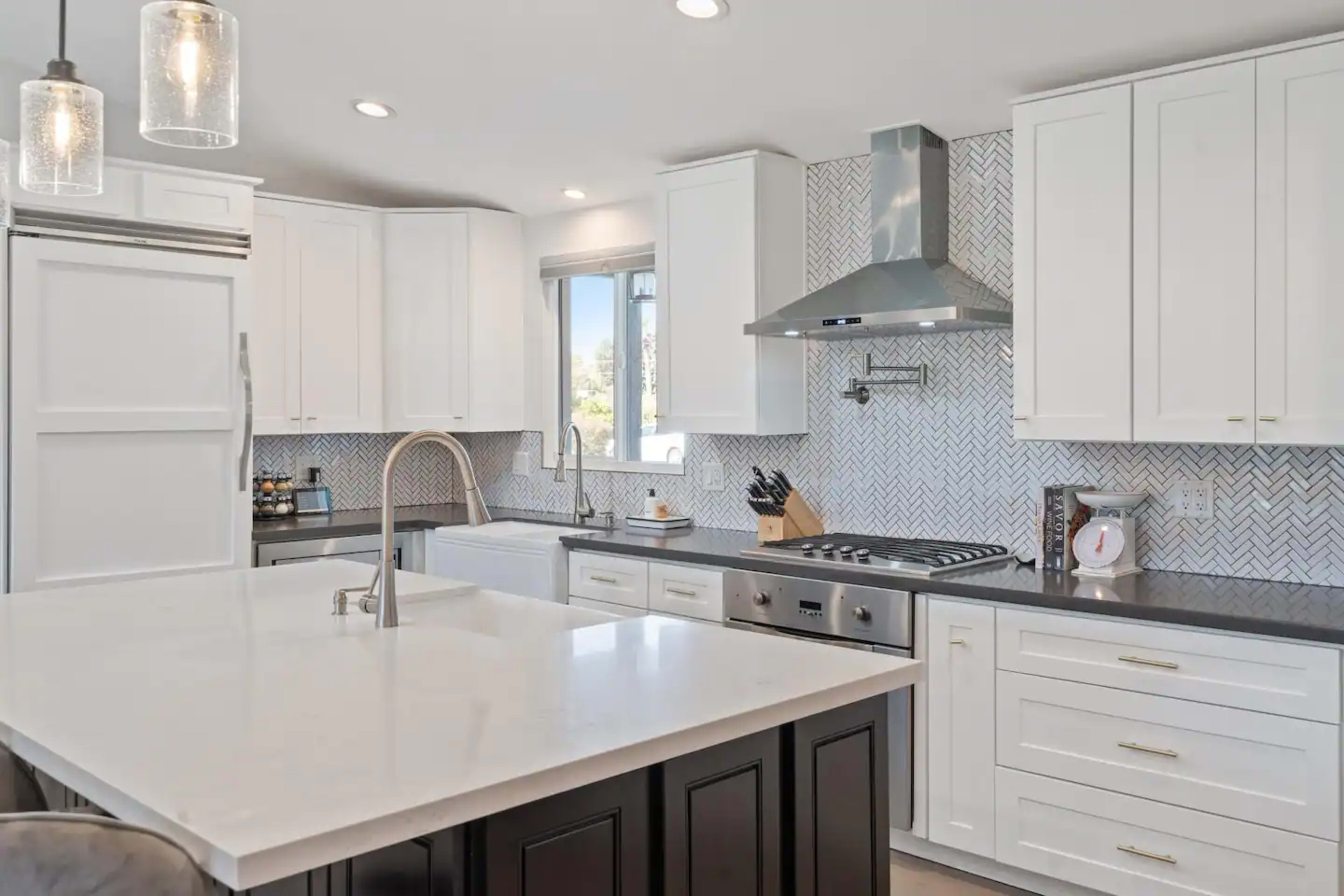 A modern kitchen features white cabinetry, a gray herringbone tiled backsplash, and a large central island with a sink.