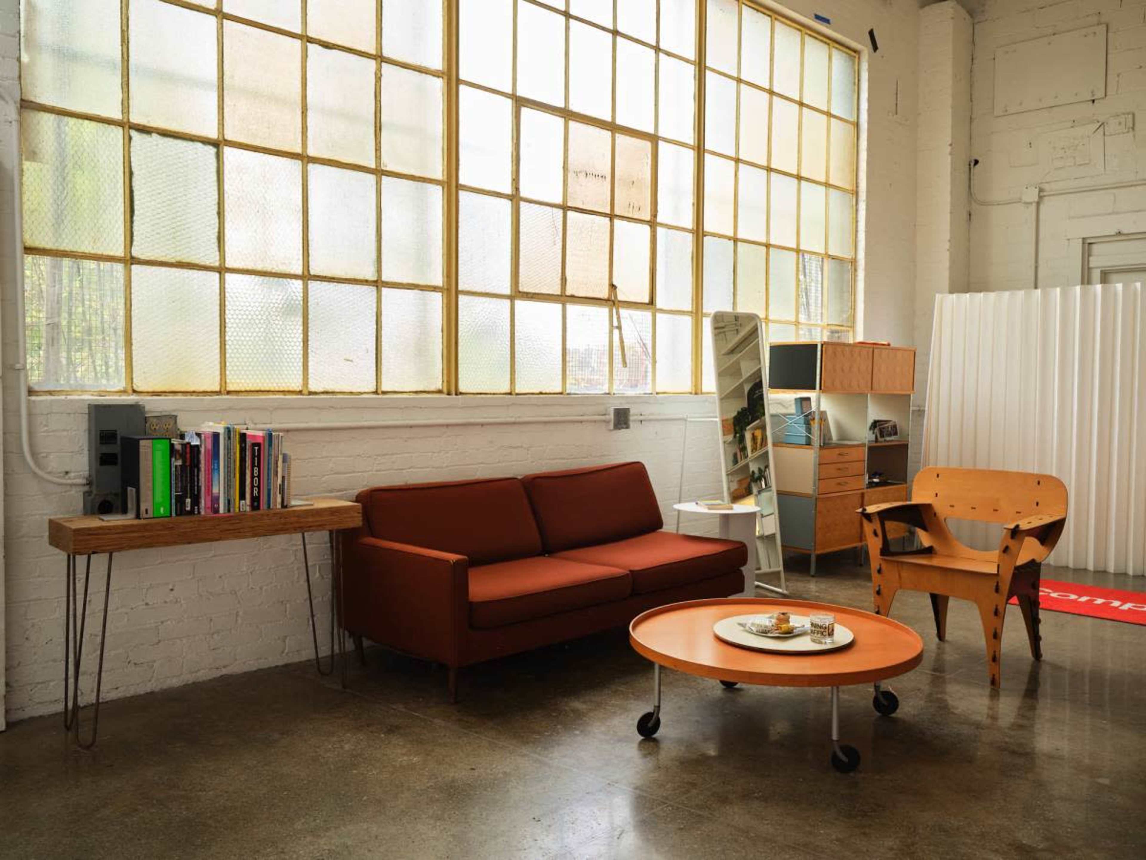 The image shows a modern living space featuring a brown sofa, a round coffee table with snacks, a wooden bookshelf filled with books, and a wooden chair, all situated near large windows with a view of natural light.