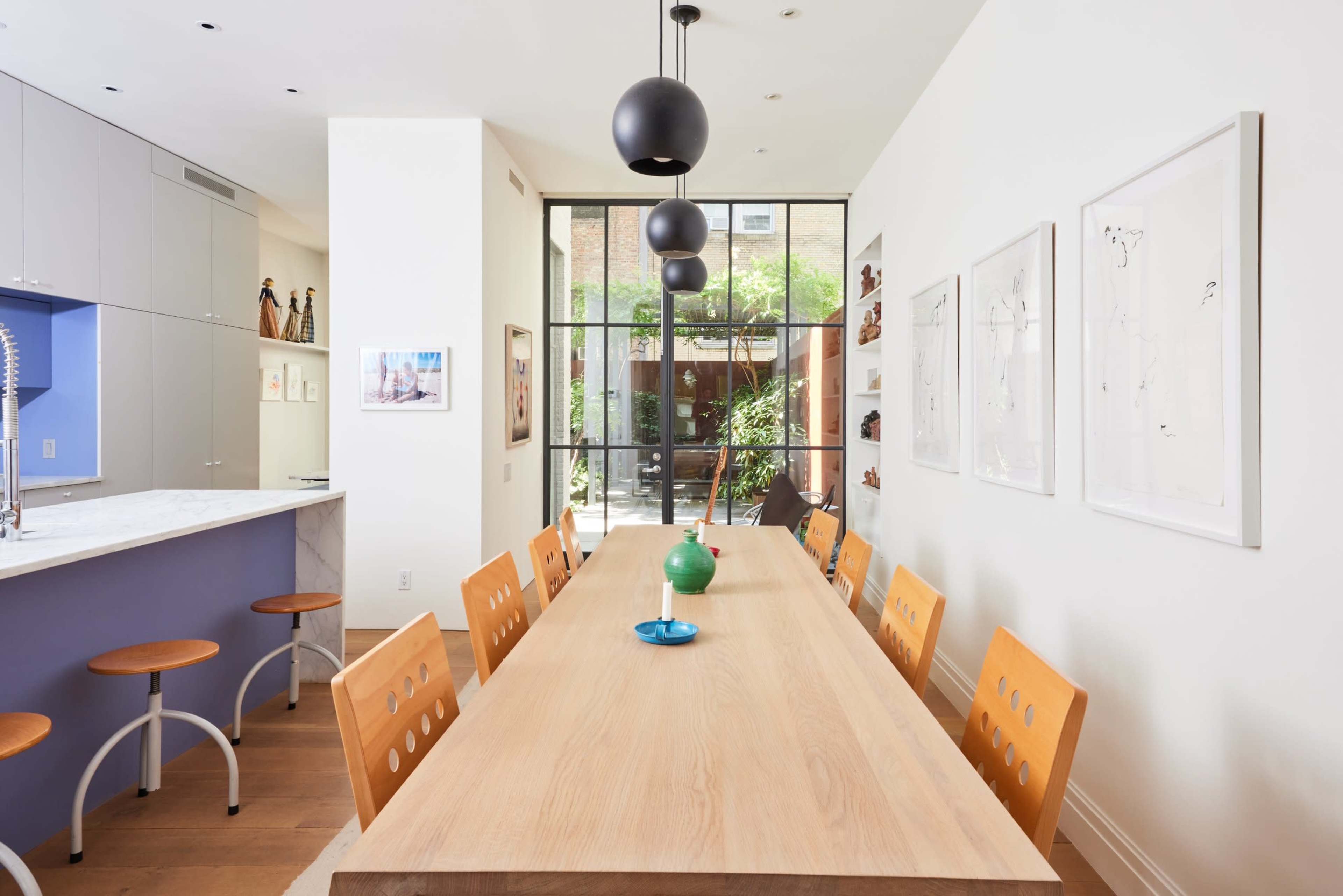 A long wooden dining table with orange chairs is set in a modern dining area featuring large windows that open to an outdoor garden.