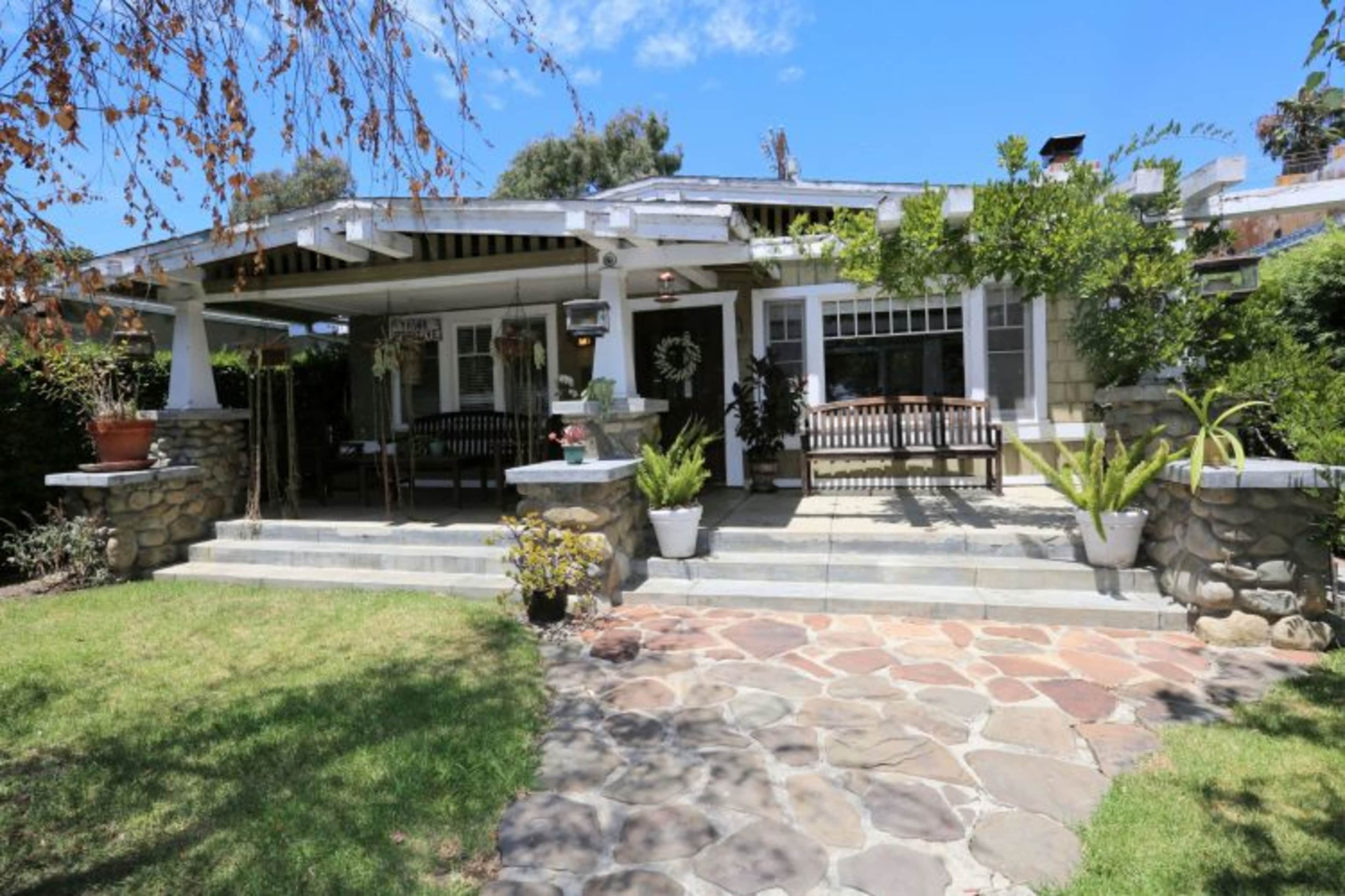 The image shows a house with a stone pathway leading to a front porch adorned with benches and potted plants.