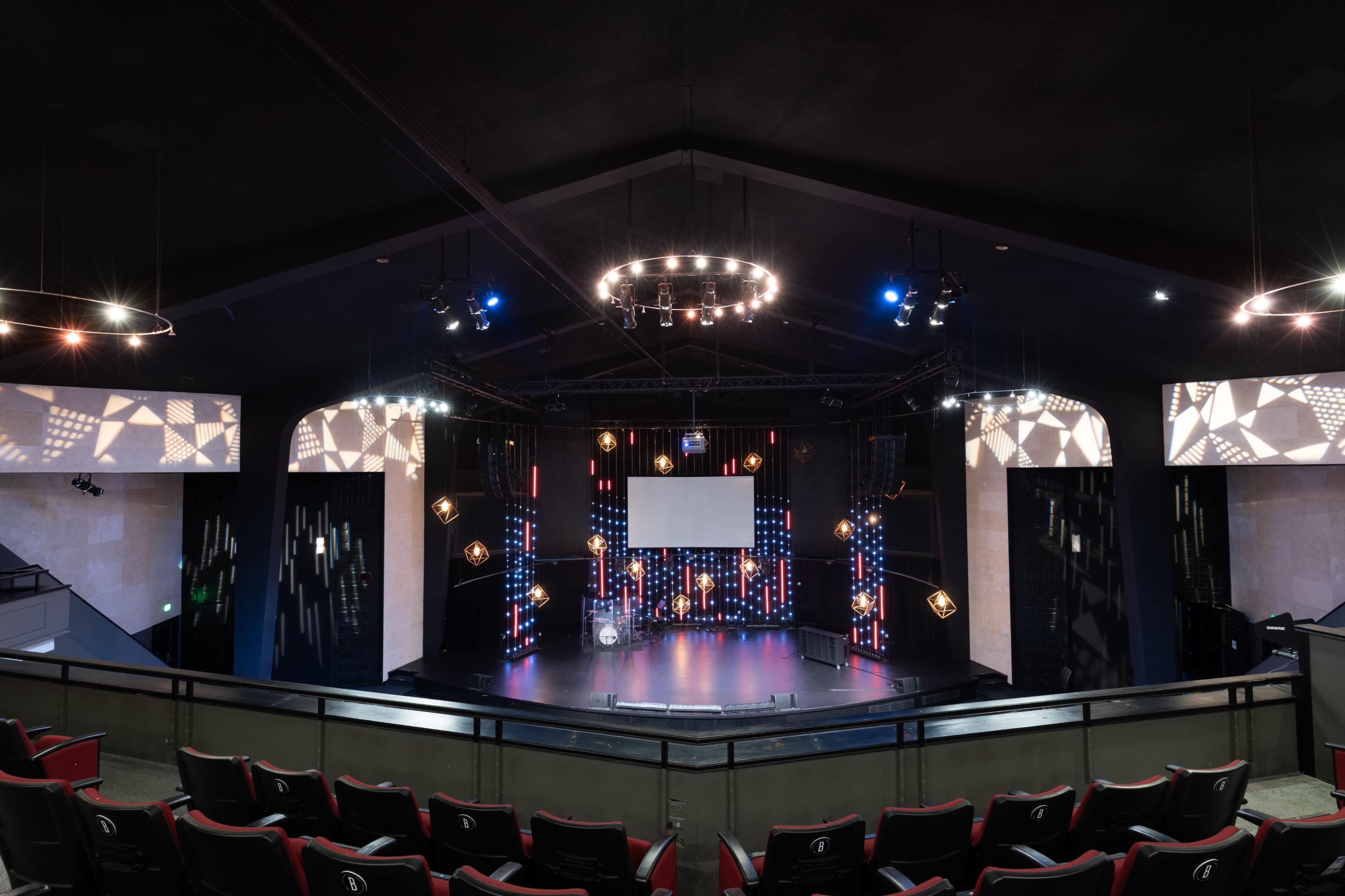 The image shows a well-lit performance stage set up in a theater, featuring lights, a large screen, and seating in the foreground.