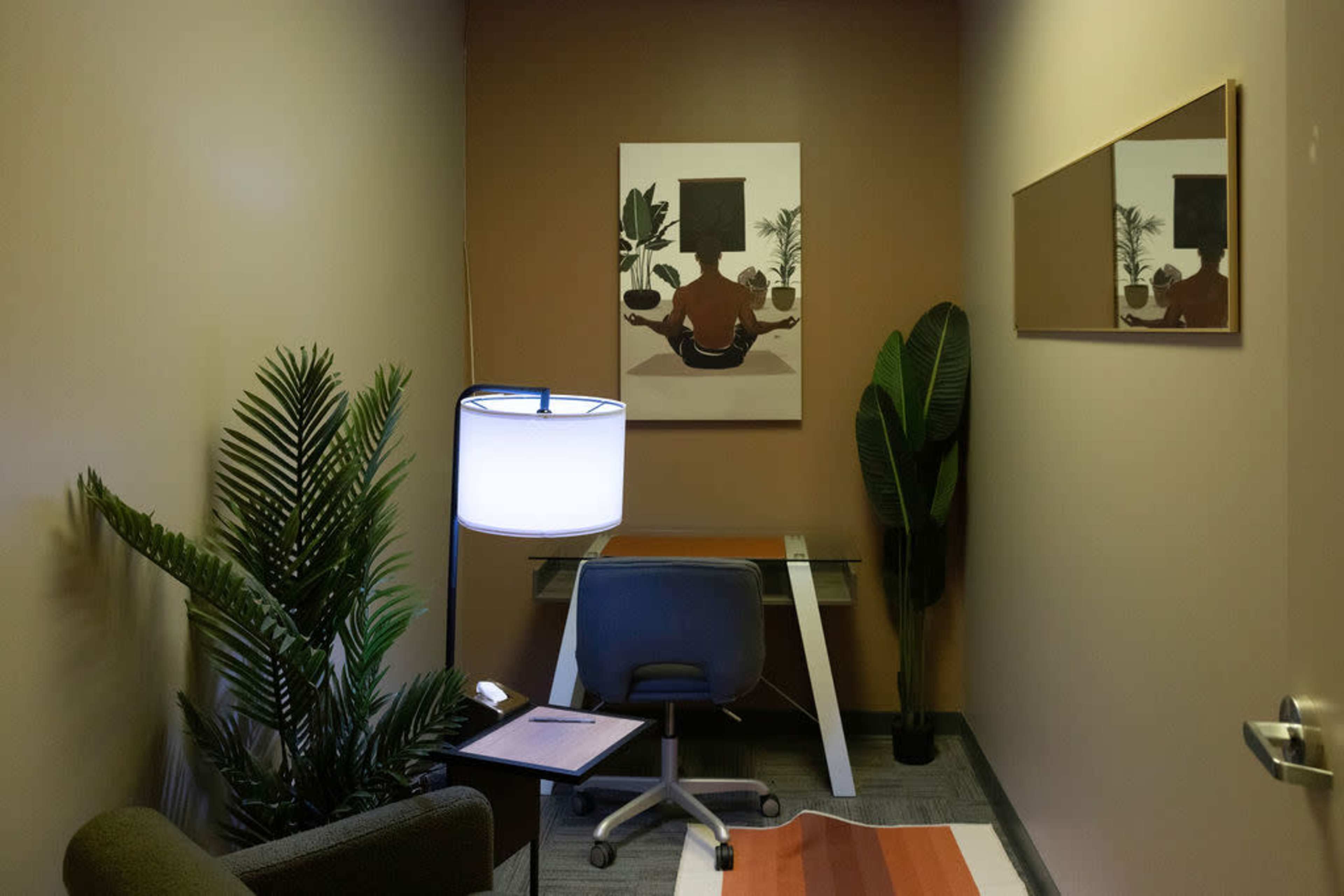 A narrow room features a desk with a chair, a lamp, and decorative plants against beige walls, with artwork hanging above the desk.
