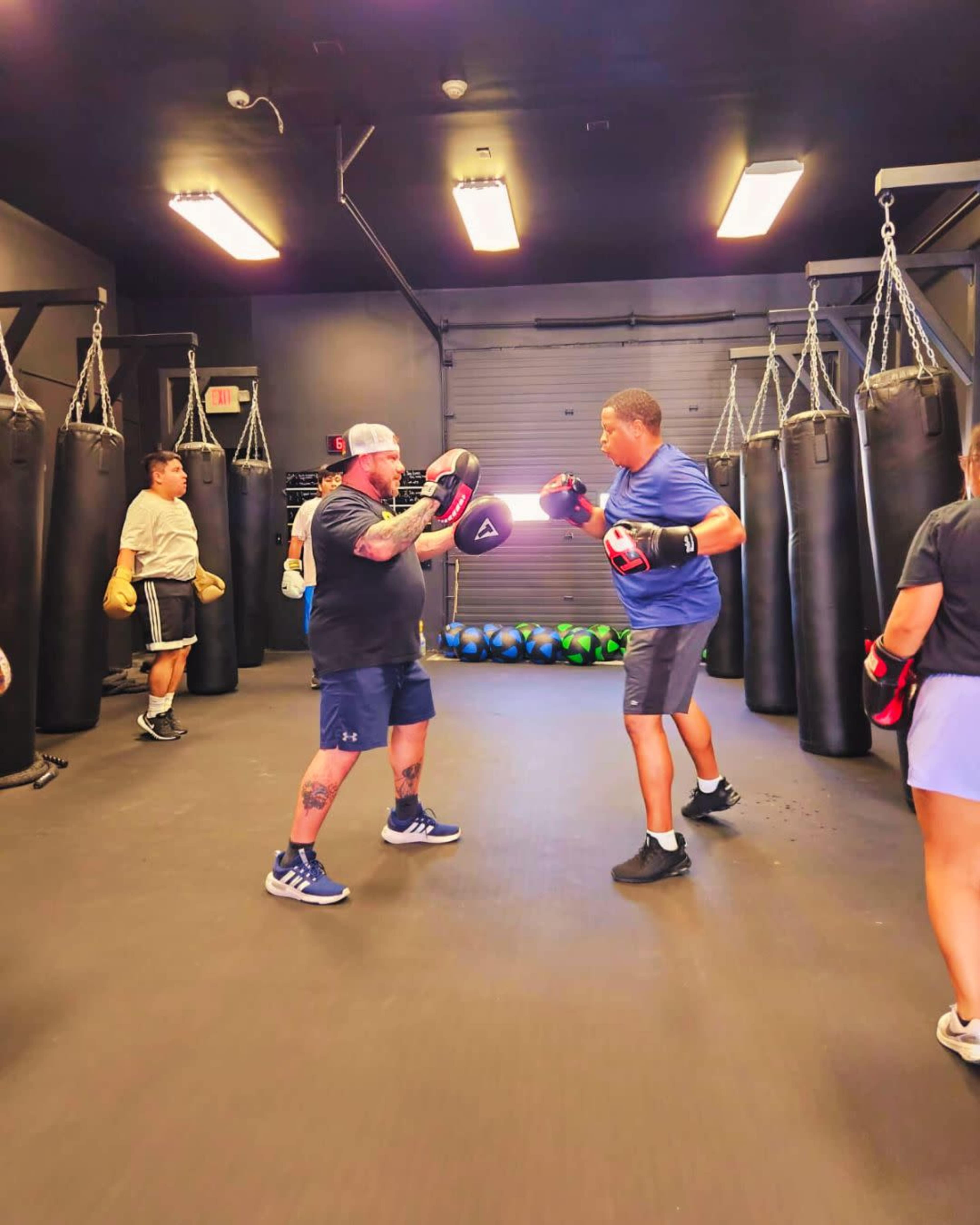 A group of individuals is engaged in a boxing training session inside a gym, with several people practicing drills with punching bags and focus mitts.