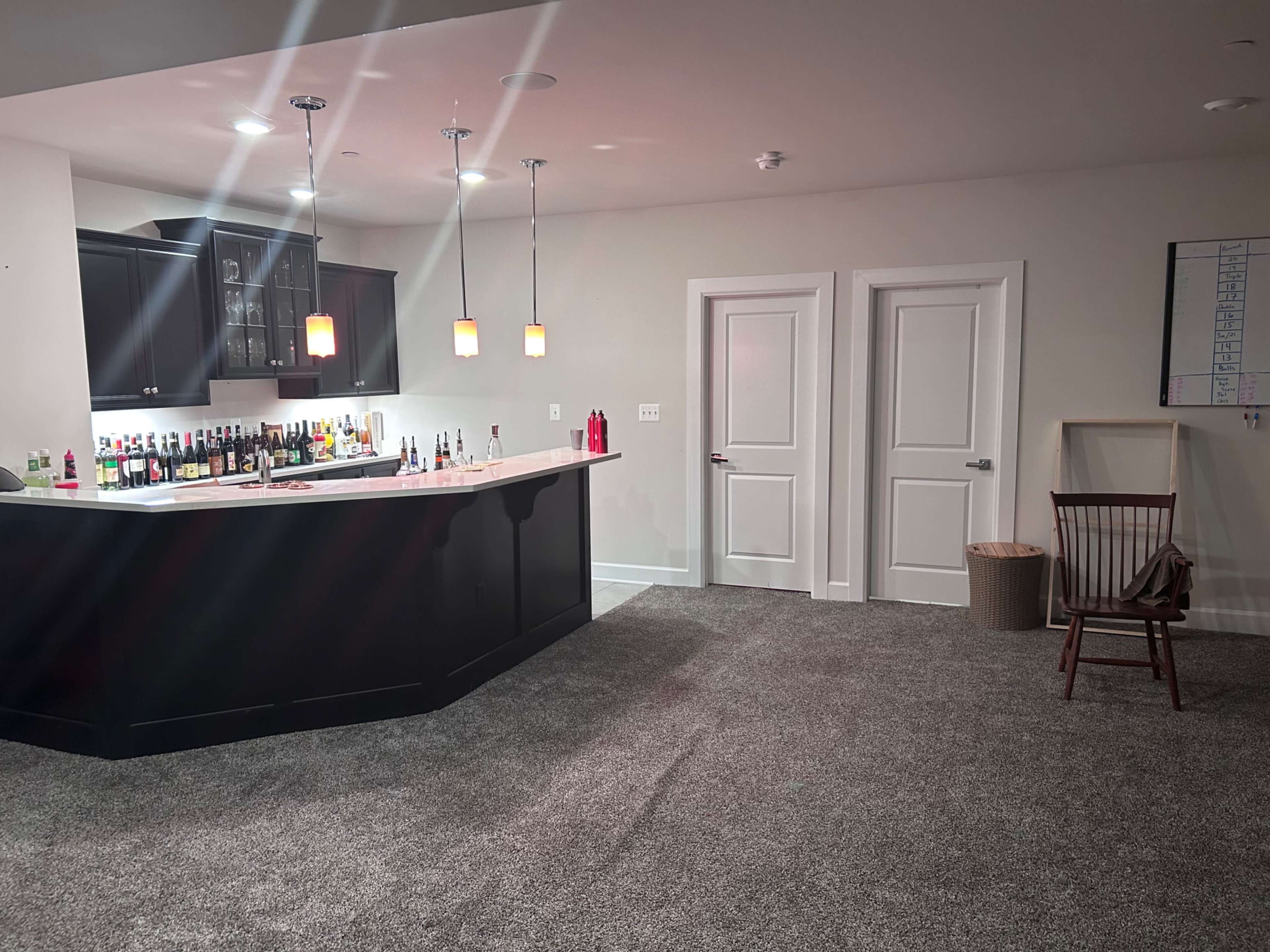 The image shows a home bar area with a curved counter, shelves filled with bottles, and two doors leading to separate rooms in a carpeted space.