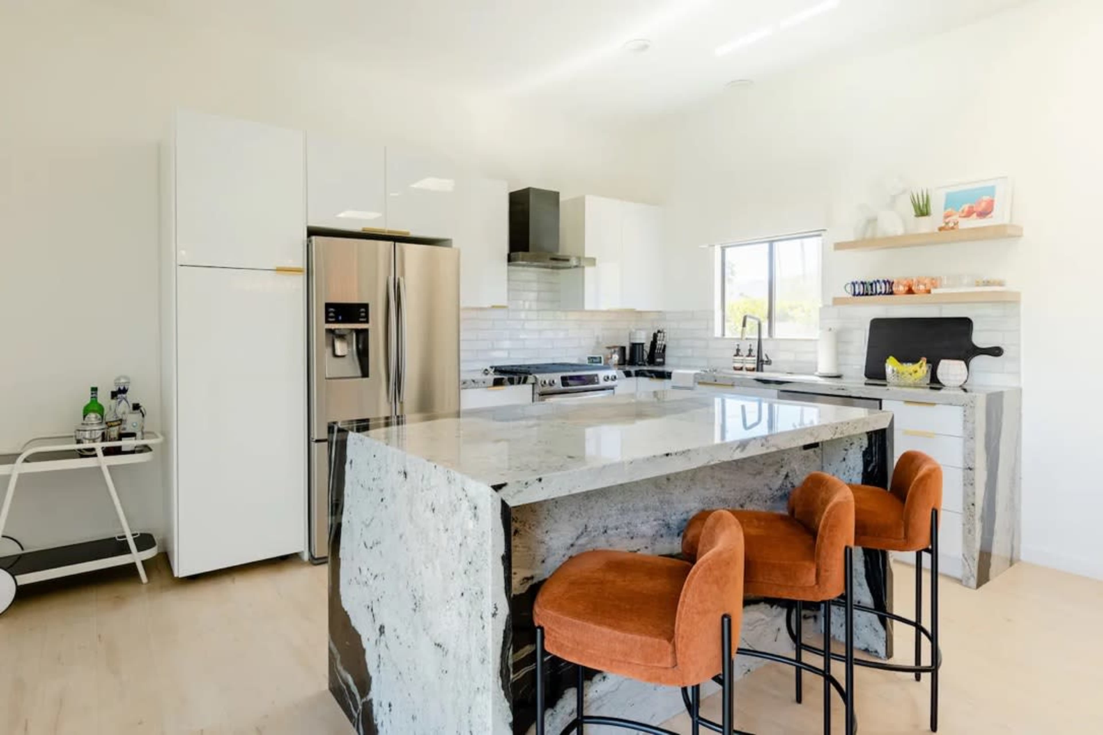 The image shows a modern kitchen with a large island featuring a speckled granite countertop, white cabinets, and orange bar stools.