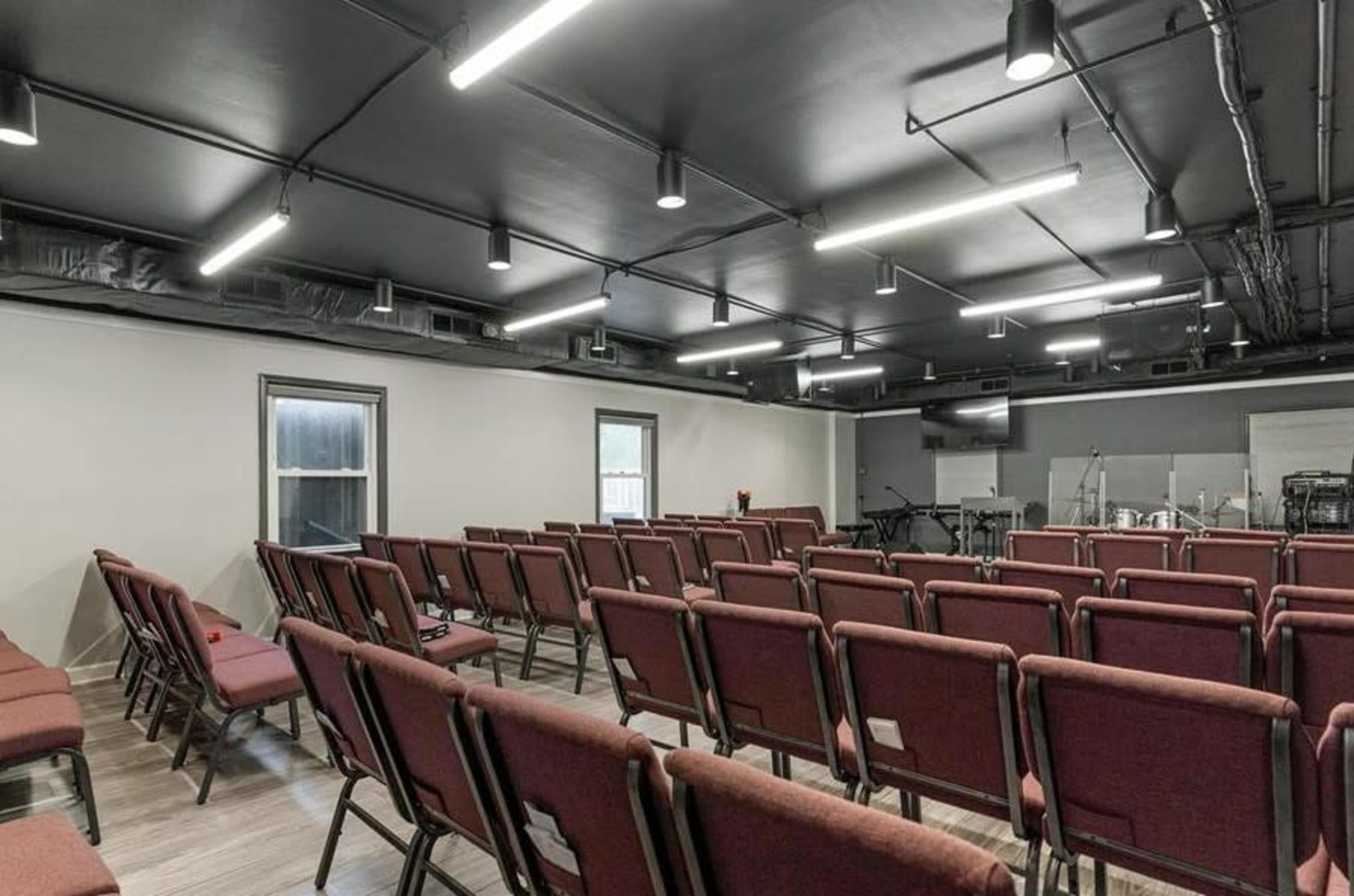 A spacious room with rows of maroon chairs facing a front stage area equipped with musical instruments and screens.