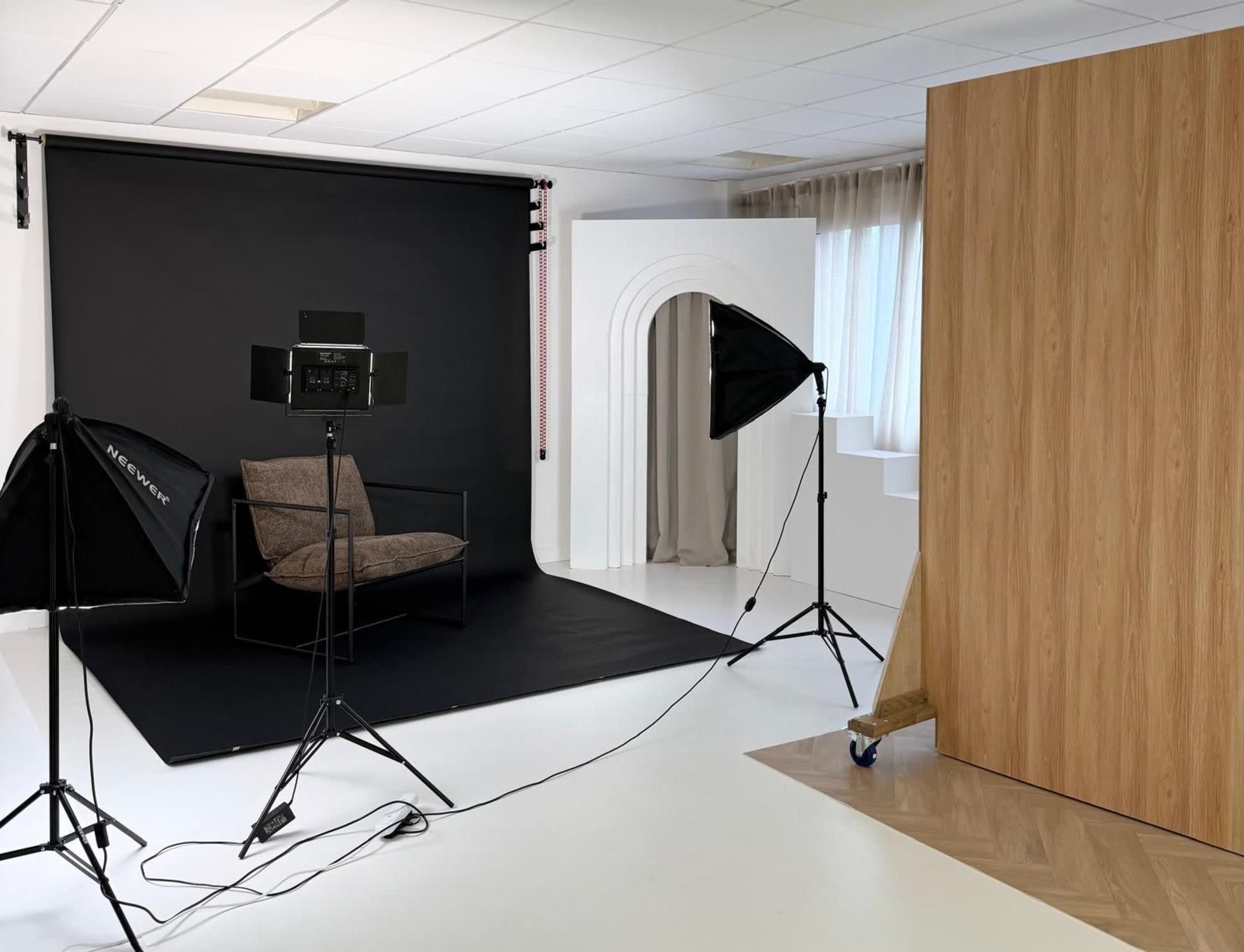The image shows a photography studio setup with a black backdrop, a chair, and lighting equipment arranged around a light wooden partition.
