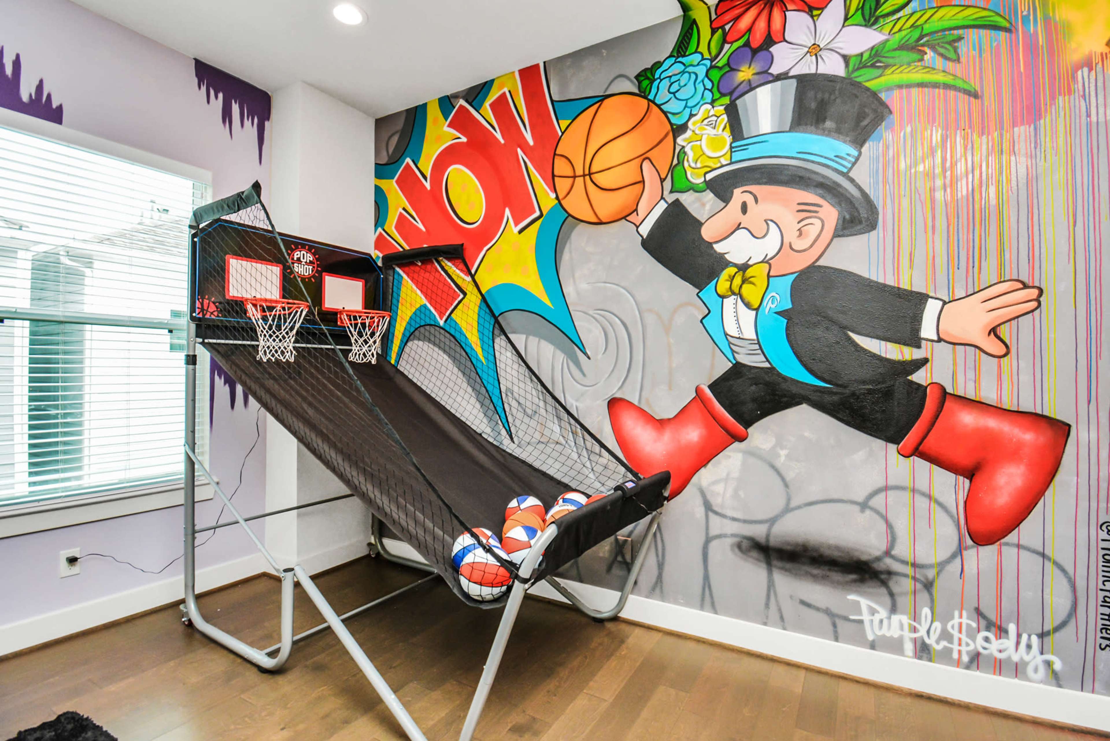 A basketball arcade game is positioned against a colorful mural featuring a character in a top hat, with basketballs piled at the base.