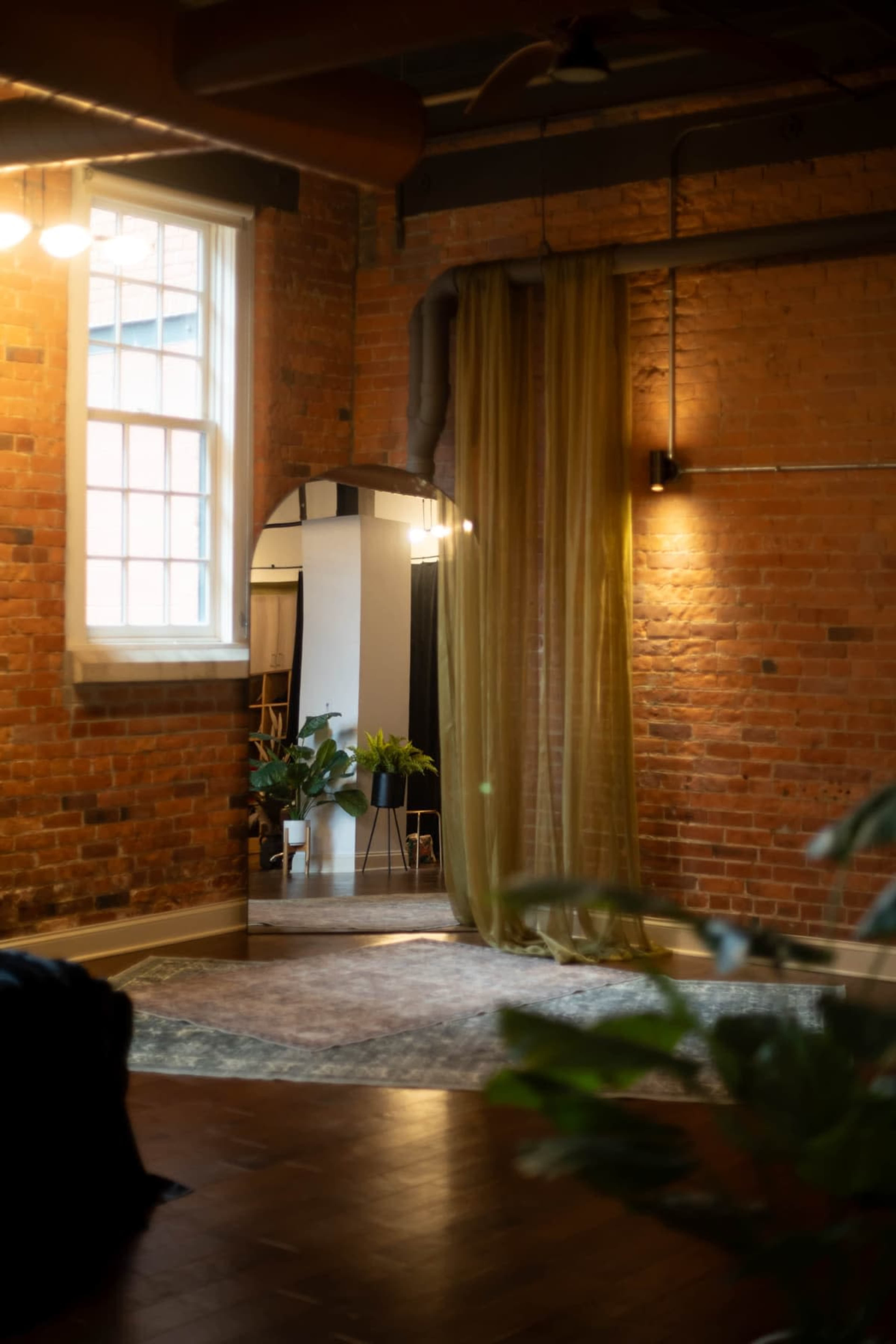 The image shows a warm, industrial-style room with exposed brick walls, a large mirror, soft lighting, and green plants integrated into the decor.